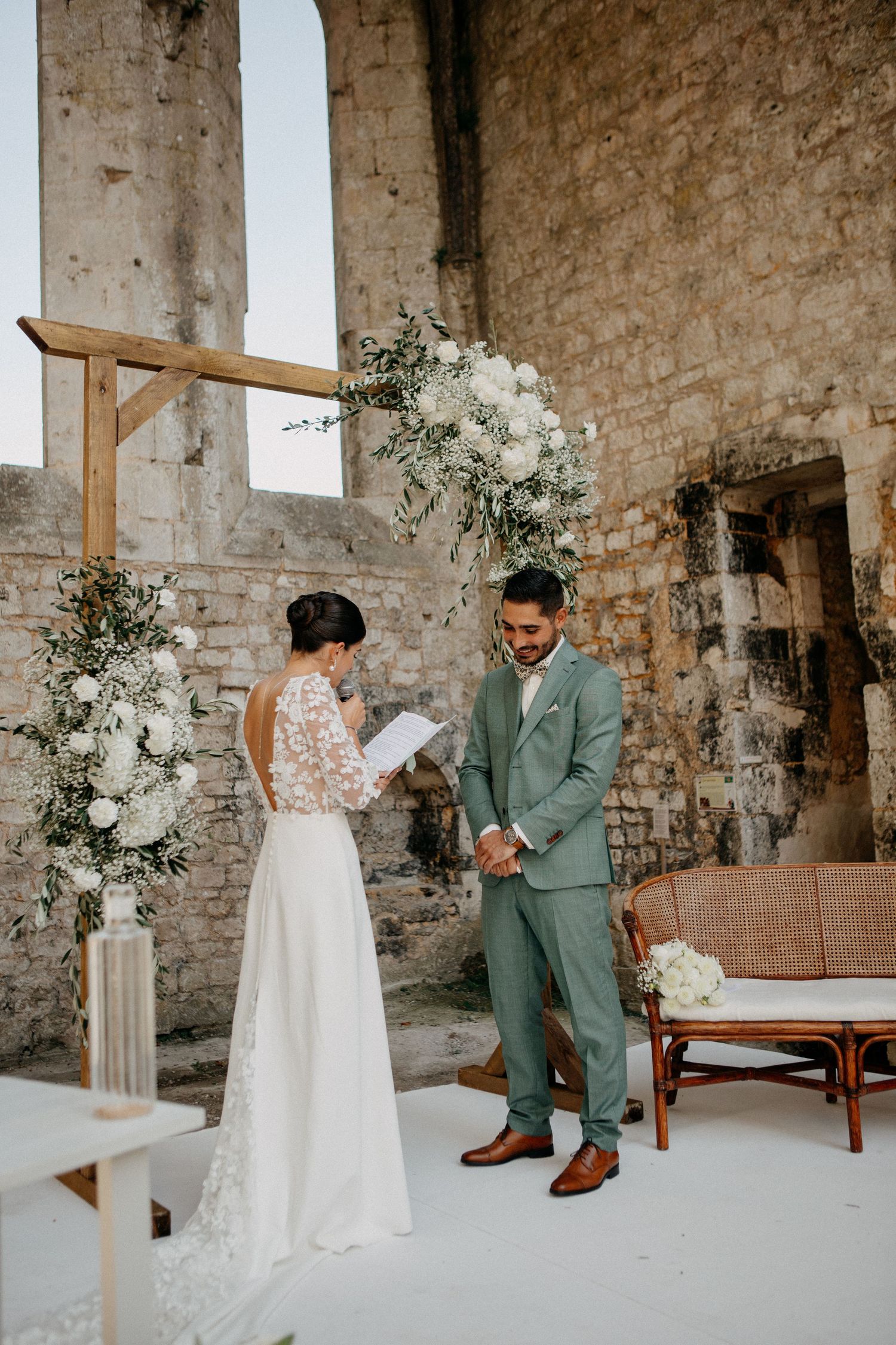 Bride reads vows to smiling groom at rustic outdoor wedding ceremony set within historic stone ruins.