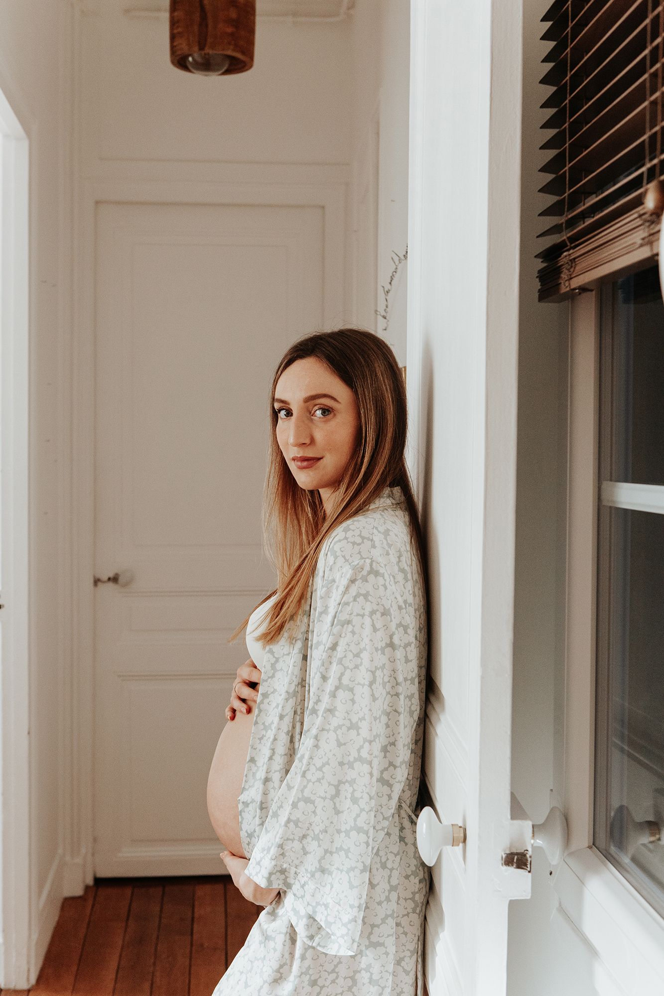 Pregnant woman in floral dress cradling baby bump while leaning against white wall near window.