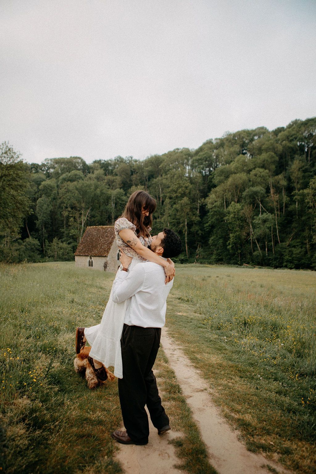 Groom lifting bride joyfully on a meadow path with a stone cottage and forest backdrop.