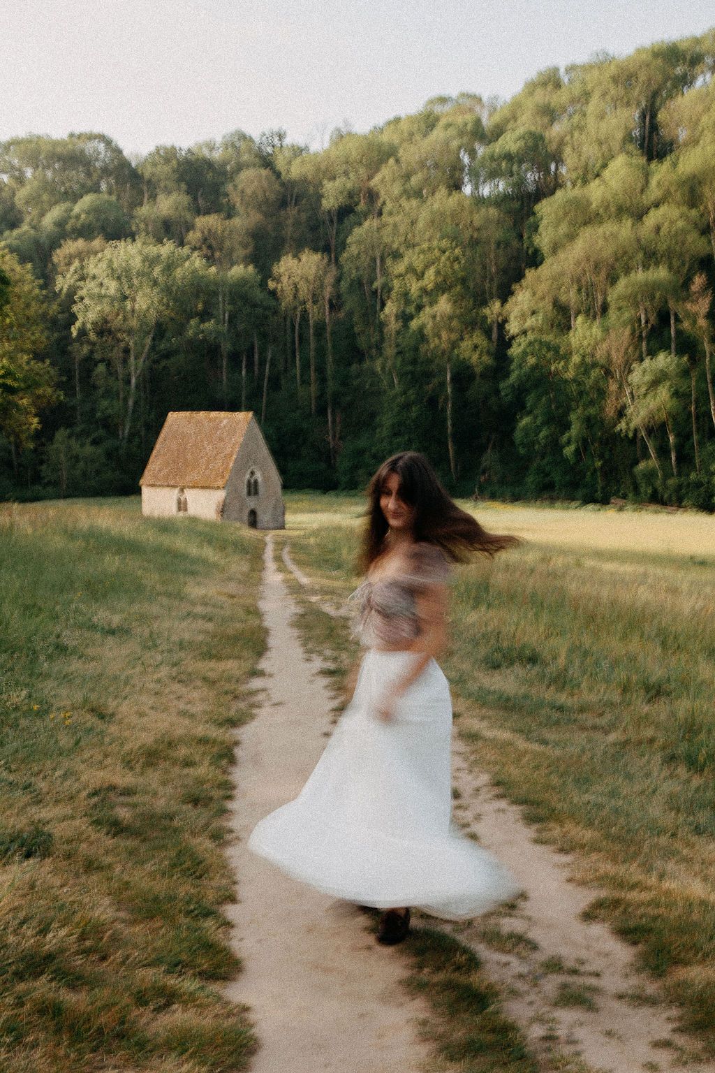 A woman in a white skirt twirls on a dirt path in a lush green meadow with a stone chapel in the background.