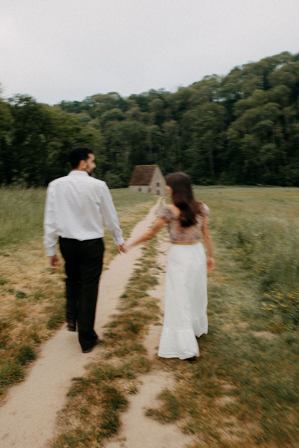 Bride and groom holding hands walking toward a stone cottage through a green countryside field.