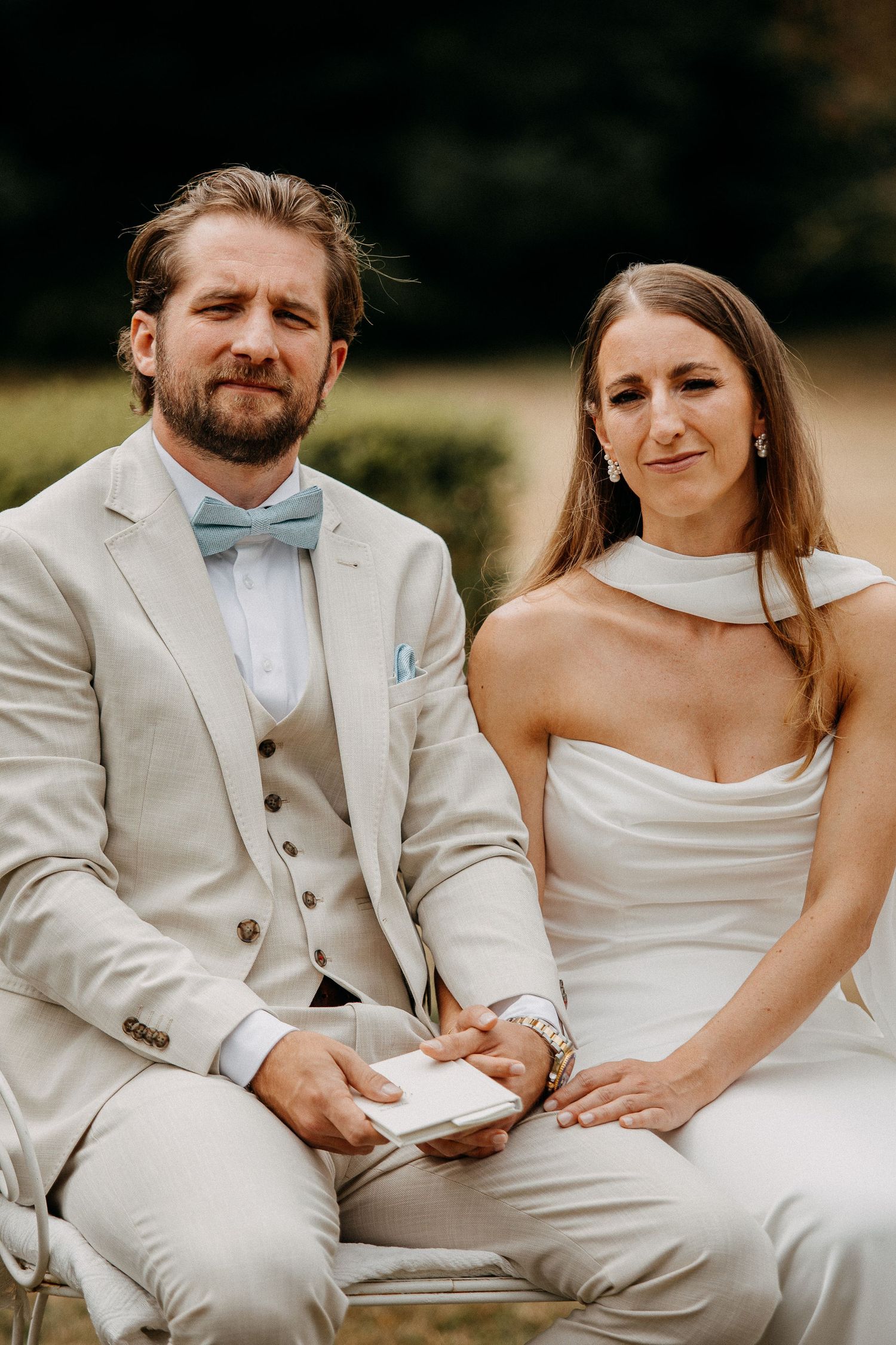 Bride in white gown and groom in beige suit with blue bow tie sit together holding hands at outdoor wedding ceremony.