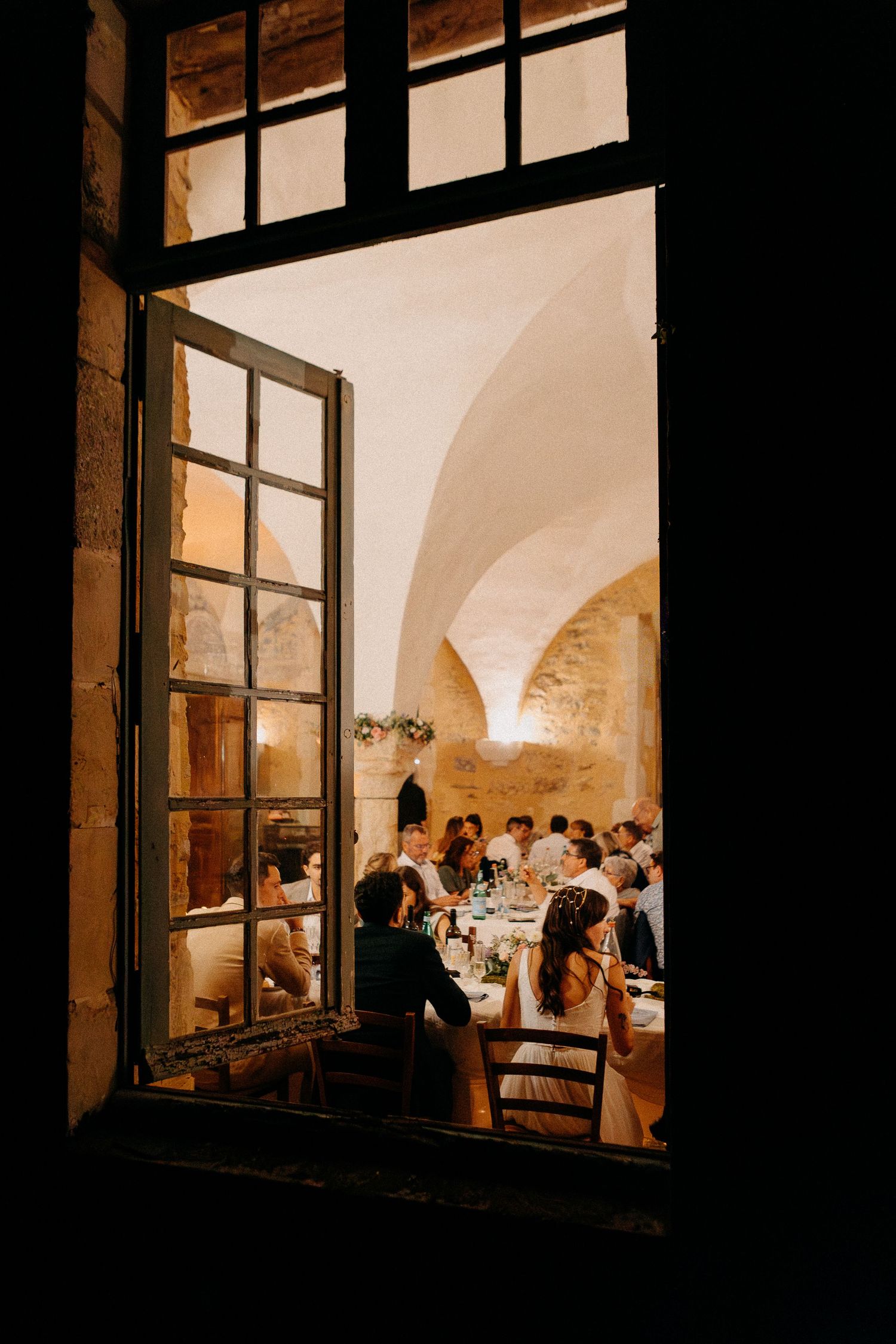 Elegant wedding reception viewed through open French doors in a stone-vaulted venue with warm candlelight.