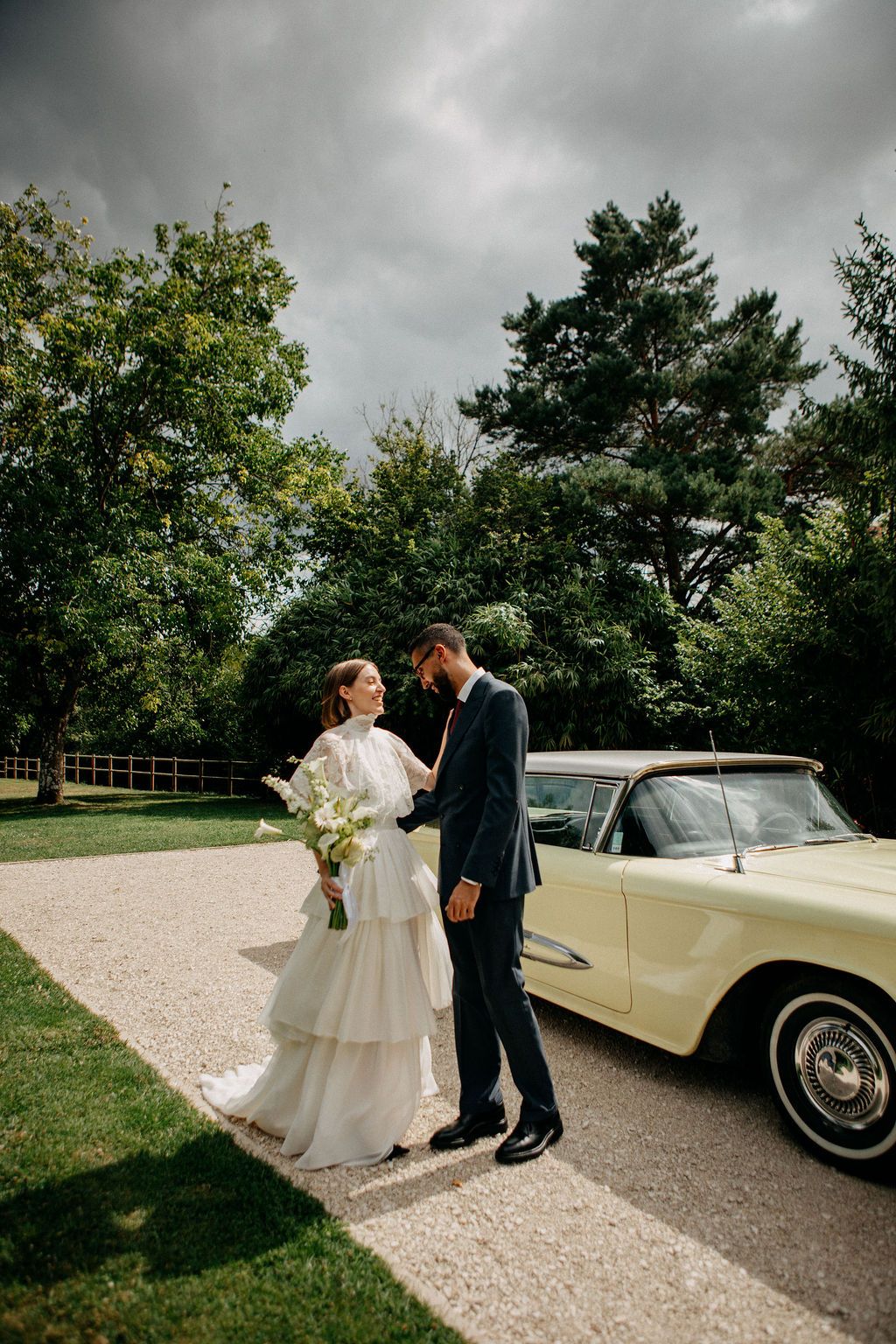 Bride and groom share an intimate moment beside a vintage yellow convertible on a gravel driveway.