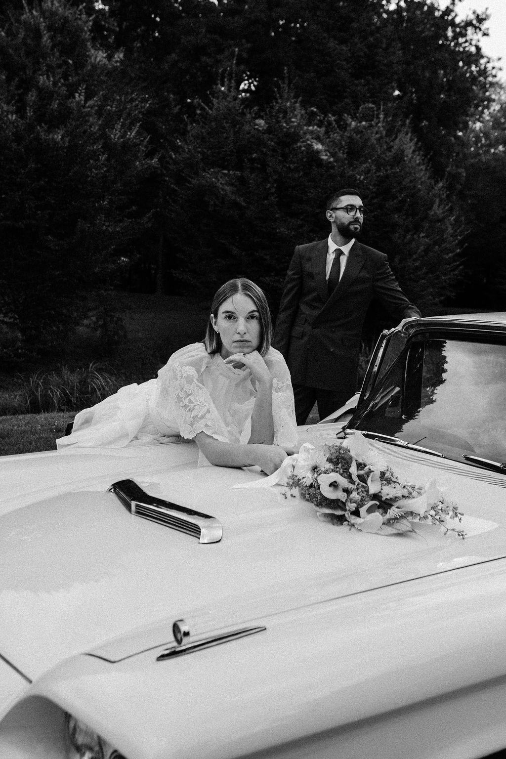 Black and white photo of bride leaning on vintage car hood with groom standing behind, wedding bouquet on car.