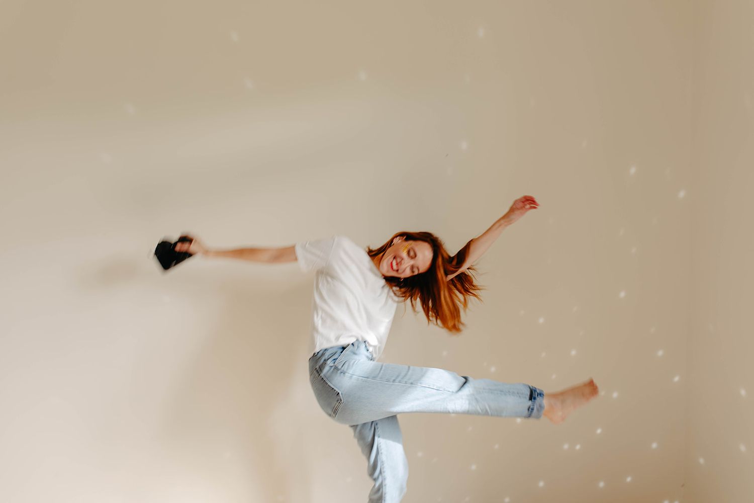 A redhead woman in a white t-shirt and jeans leaping joyfully against a light beige wall.