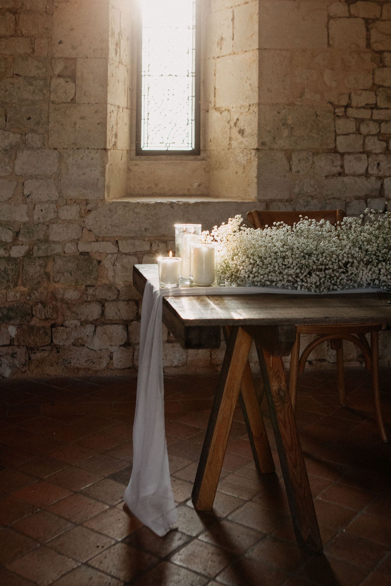 Rustic wedding altar with white candles, baby's breath flowers, and draped fabric on wooden trestle table in stone chapel.