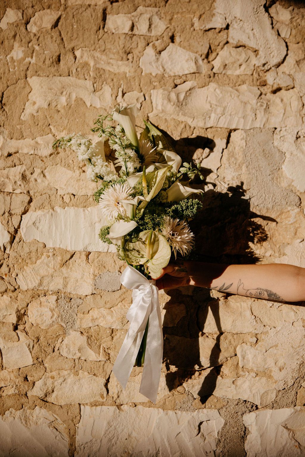 A hand holds a white wedding bouquet with calla lilies and greenery against a rustic stone wall.