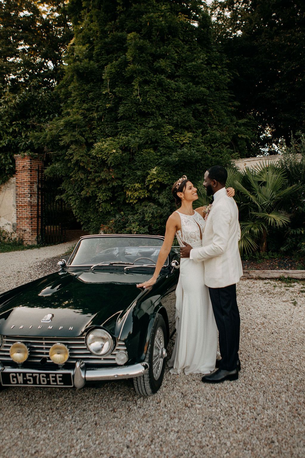 Interracial wedding couple posing beside a vintage black Triumph convertible on a gravel driveway.