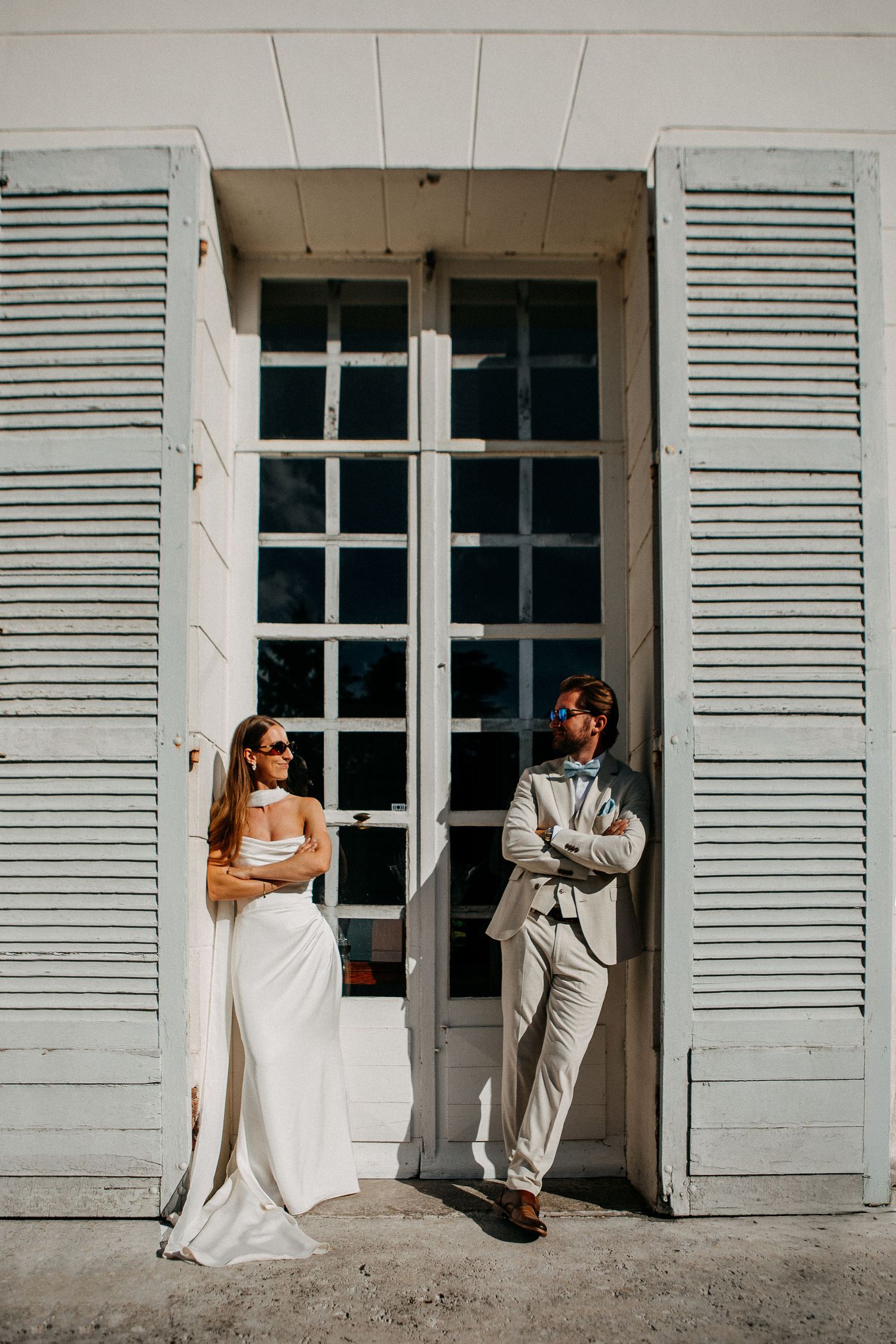 Bride in white gown and groom in tan suit lean against vintage shuttered doors in a bright outdoor wedding portrait.
