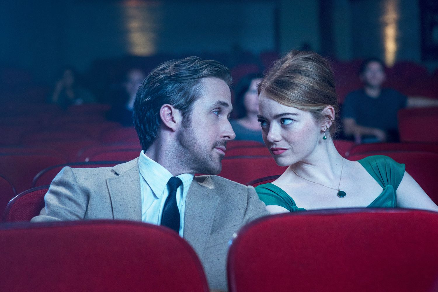 A man and woman share an intimate glance while seated in a red-seated movie theater.