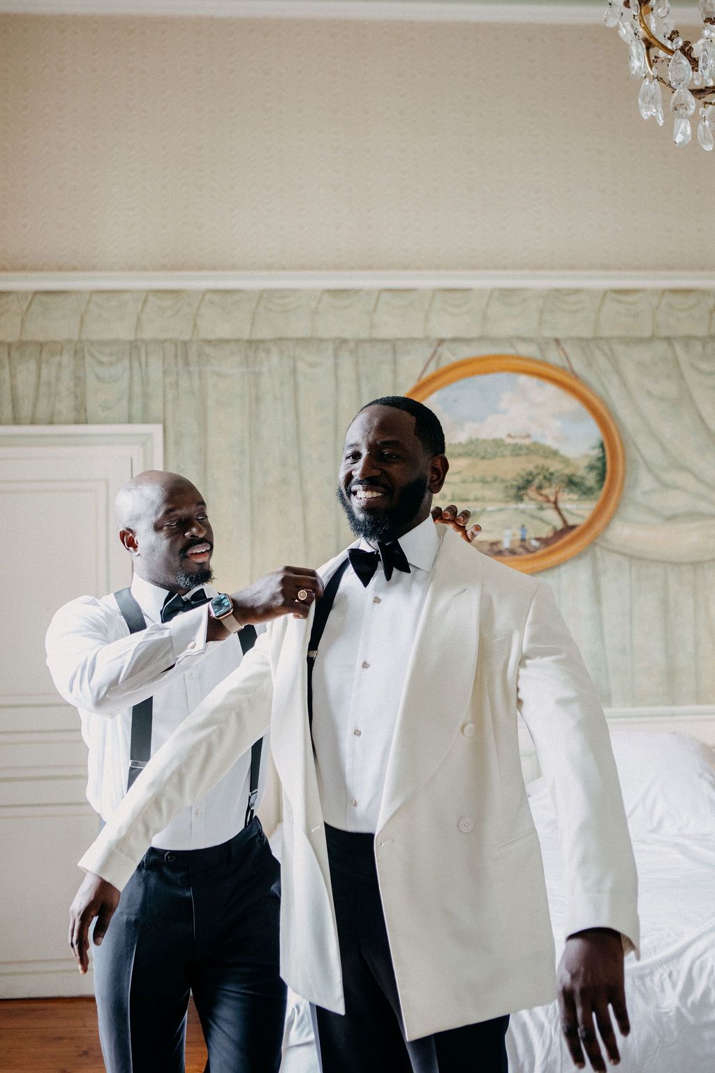 A smiling groom in a white tuxedo jacket gets his bow tie adjusted by a groomsman.