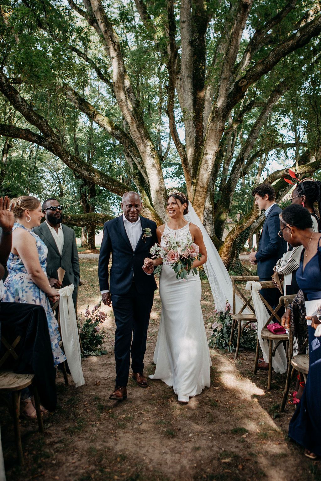 Bride in white gown walks down the aisle with escort through a sunlit outdoor woodland wedding ceremony.