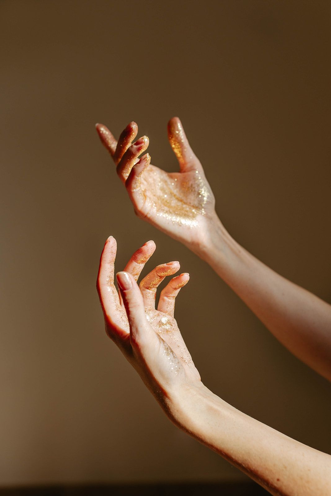 Two elegant hands with gold glitter raised gracefully against a dark beige background.