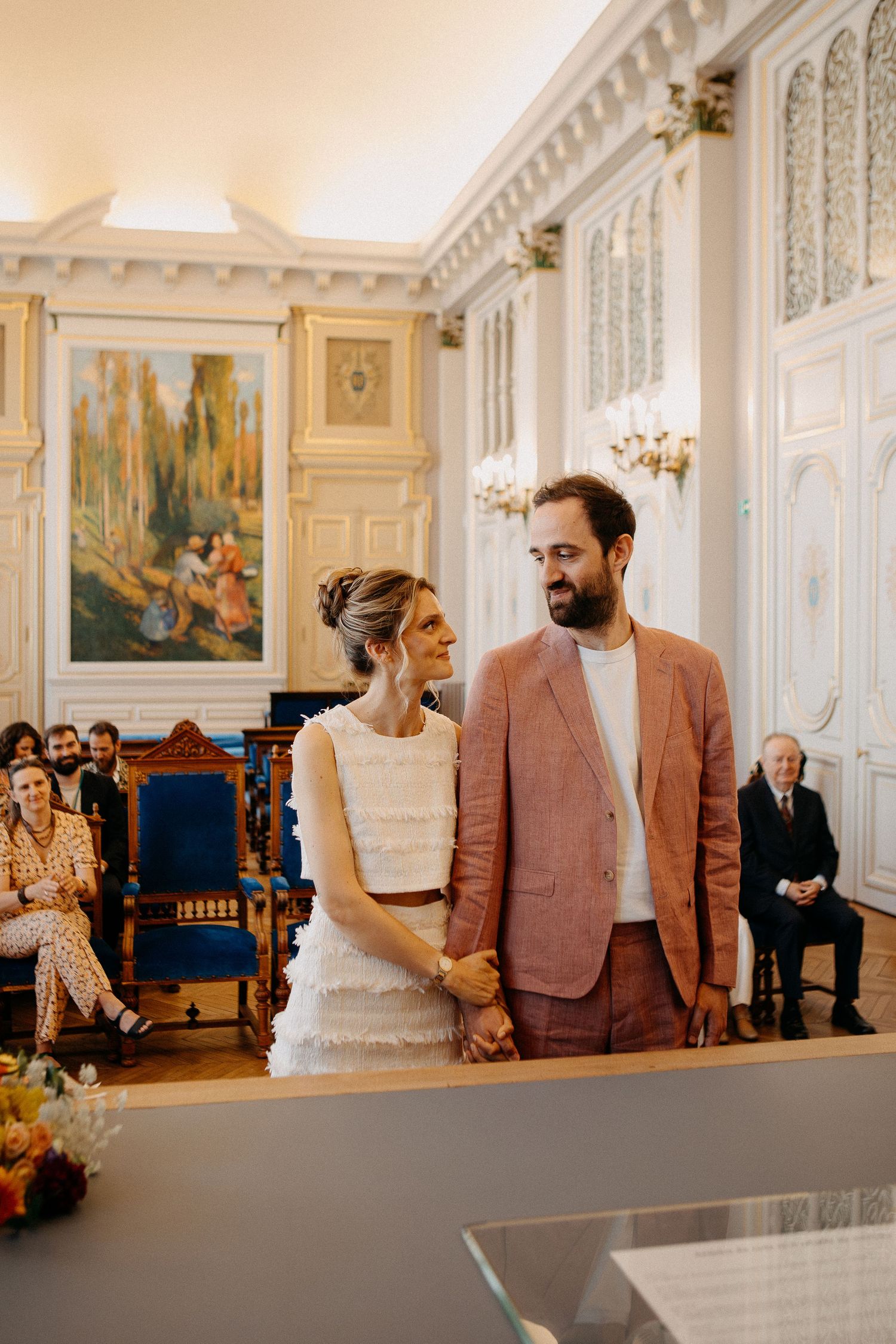 Couple holding hands at civil wedding ceremony in elegant white room with ornate decor and guests seated behind.