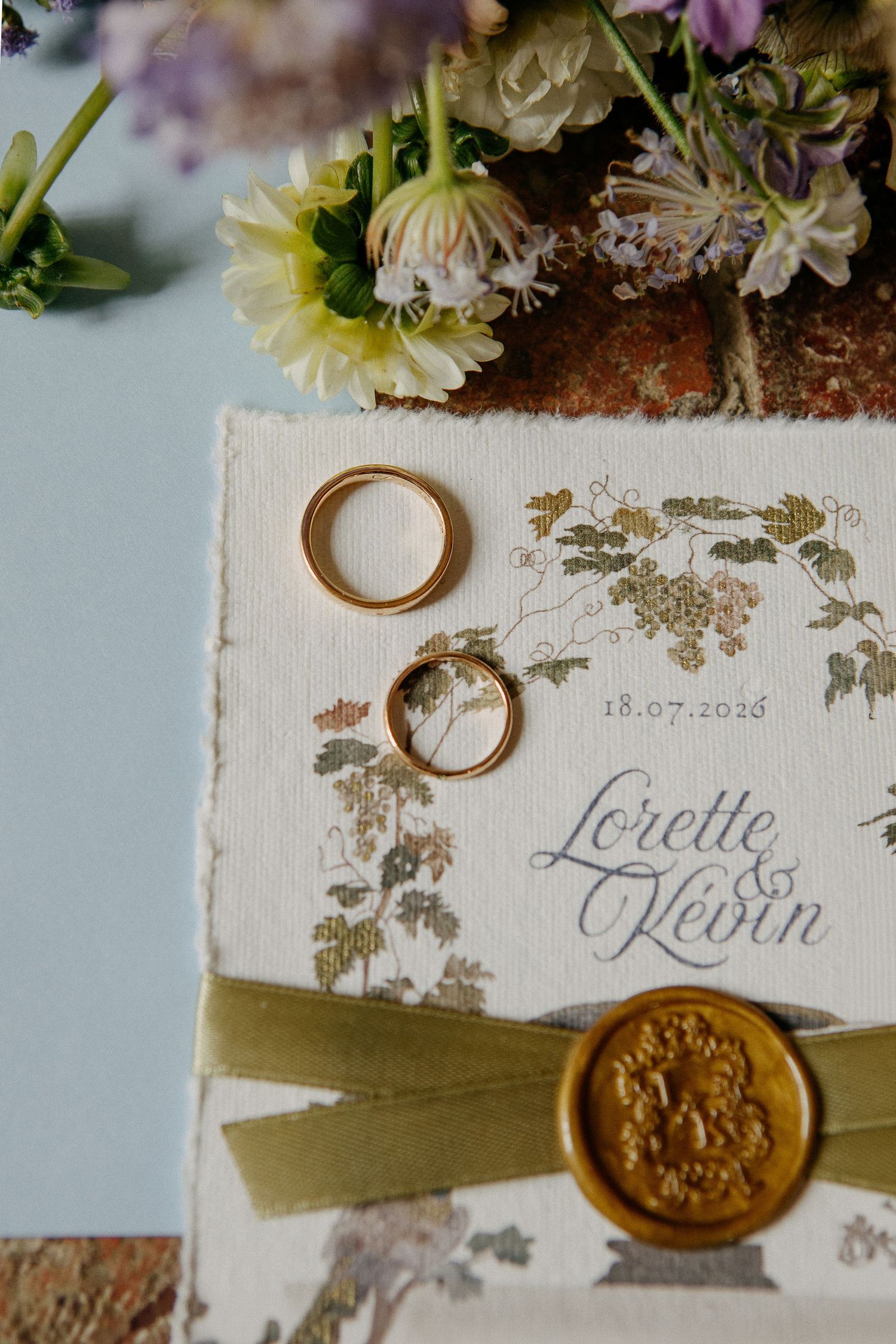 Gold wedding rings on floral invitation for Lorette and Kevin dated 18.07.2025 with wax seal and ribbon.