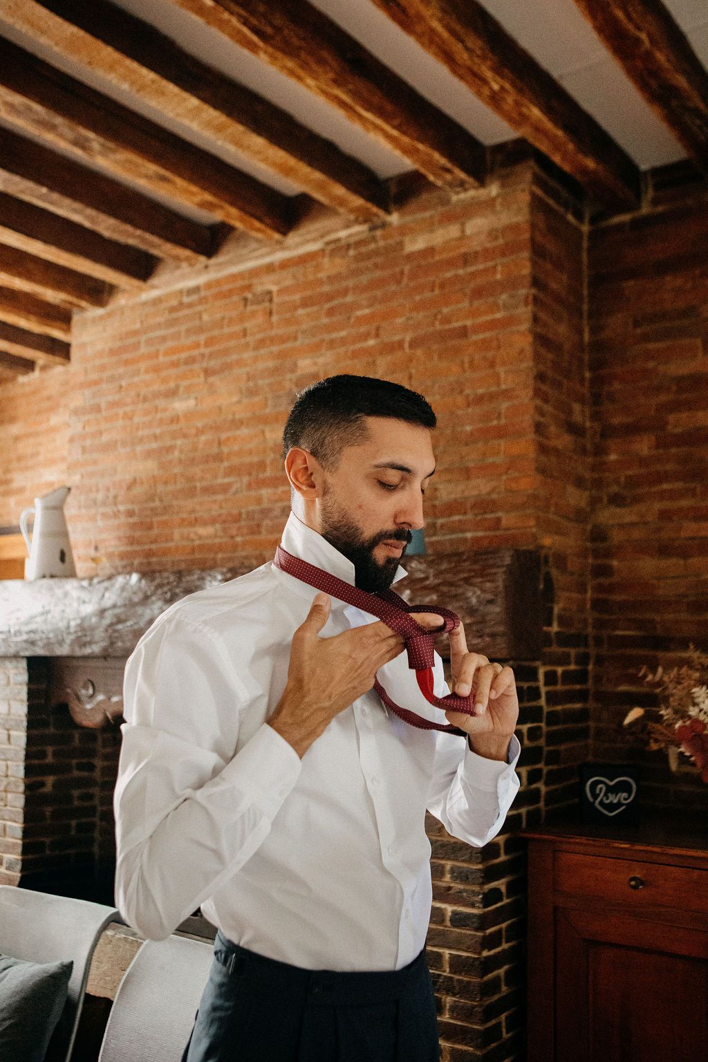 Groom in white shirt adjusting burgundy tie in rustic brick fireplace room.