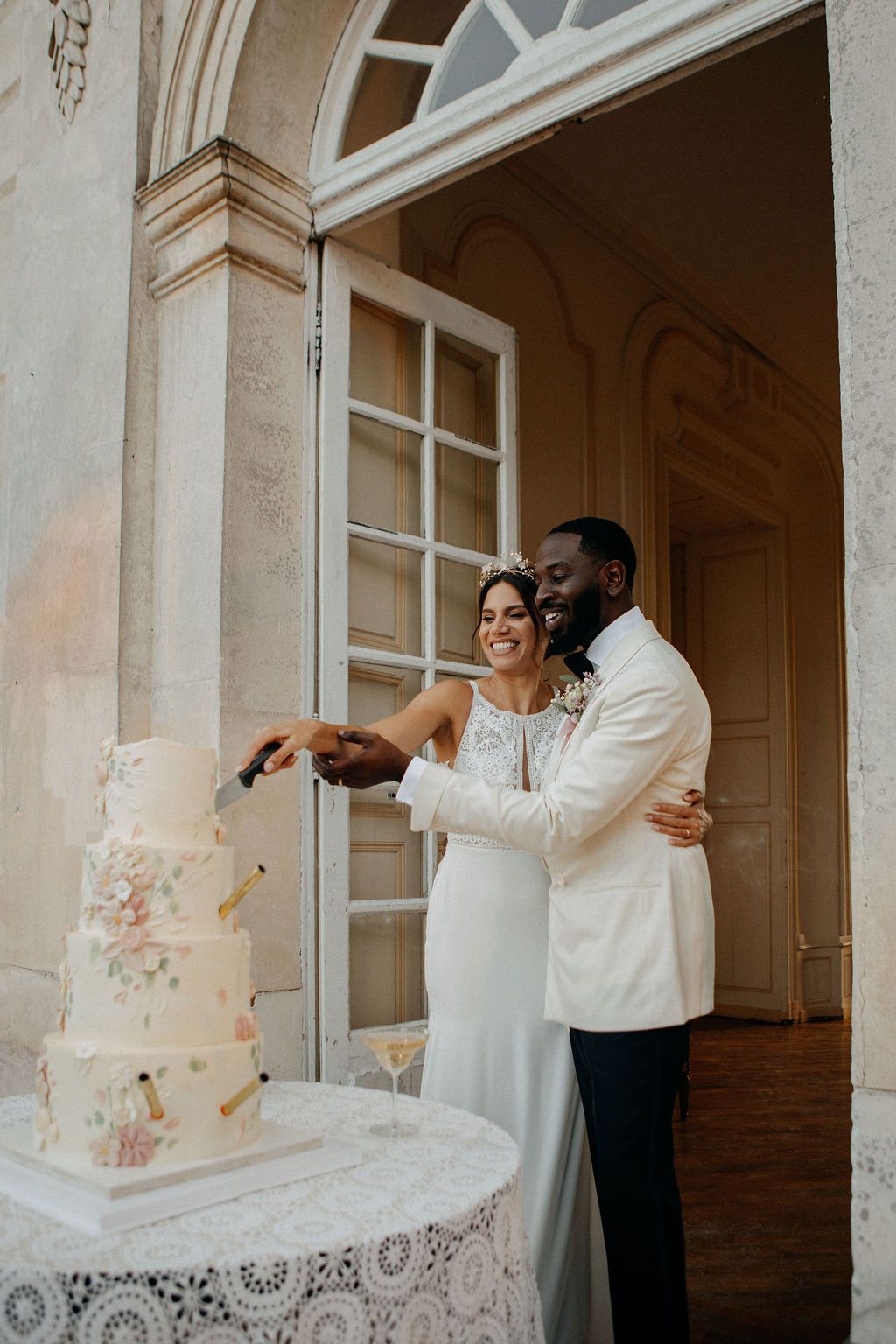 Smiling interracial couple cutting a elegant multi-tiered white wedding cake with floral details at a chateau.