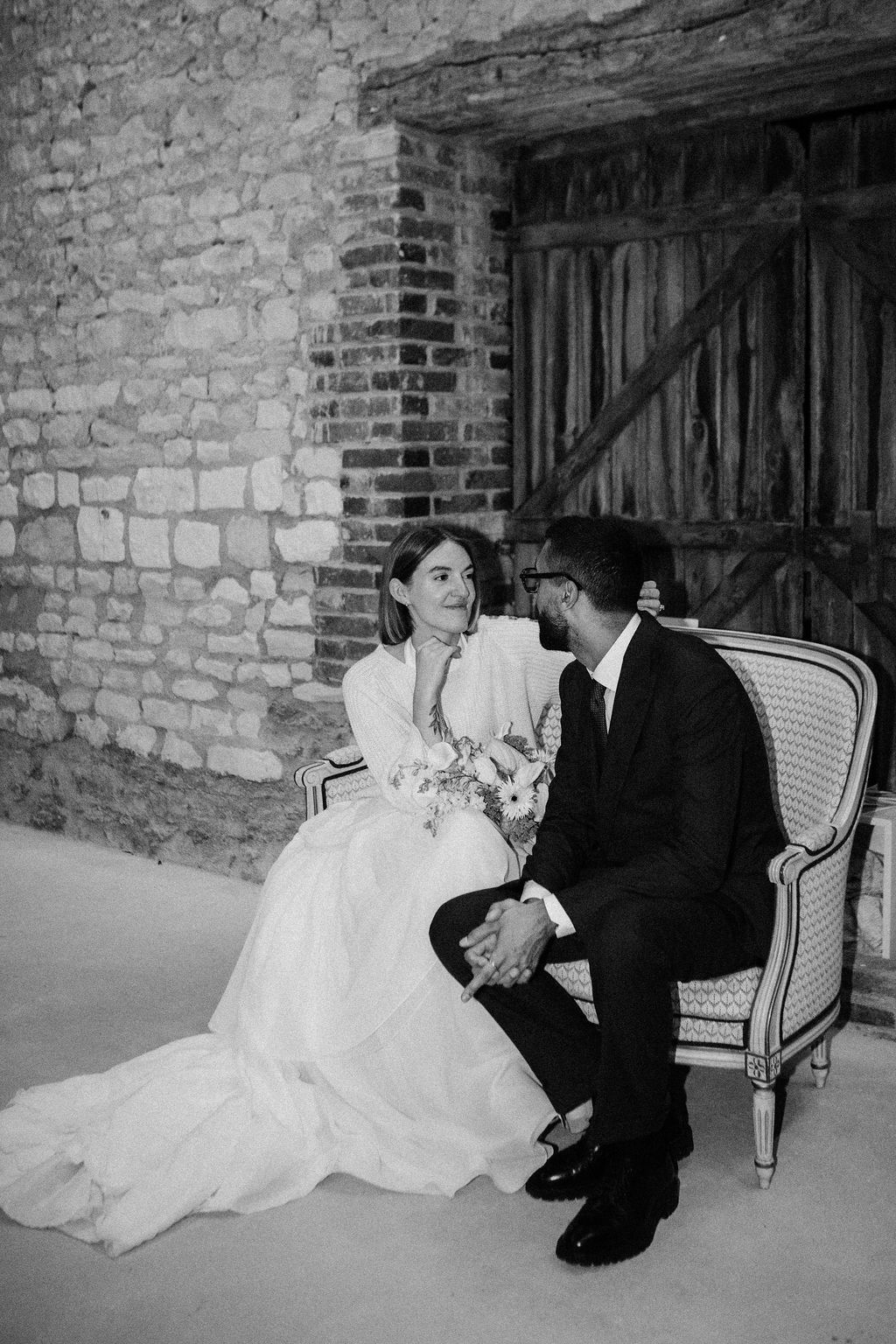 Black and white photo of bride and groom sitting together on elegant chair against rustic stone wall.