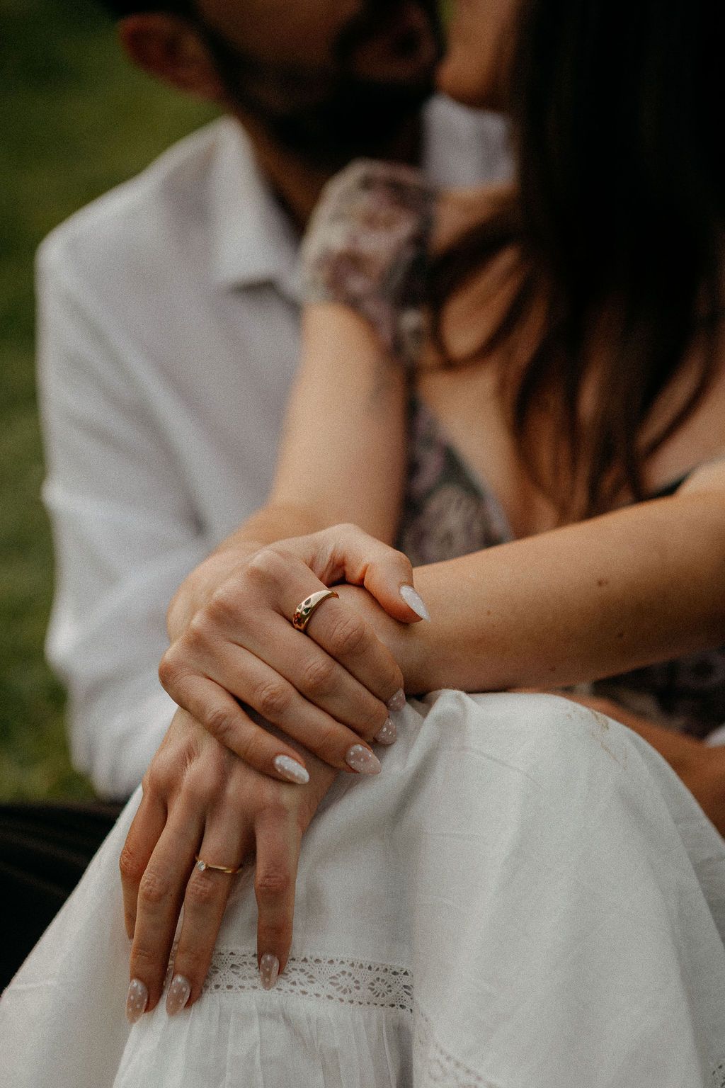 Close-up of engaged couple's hands showing gold rings, woman in white dress with floral top.