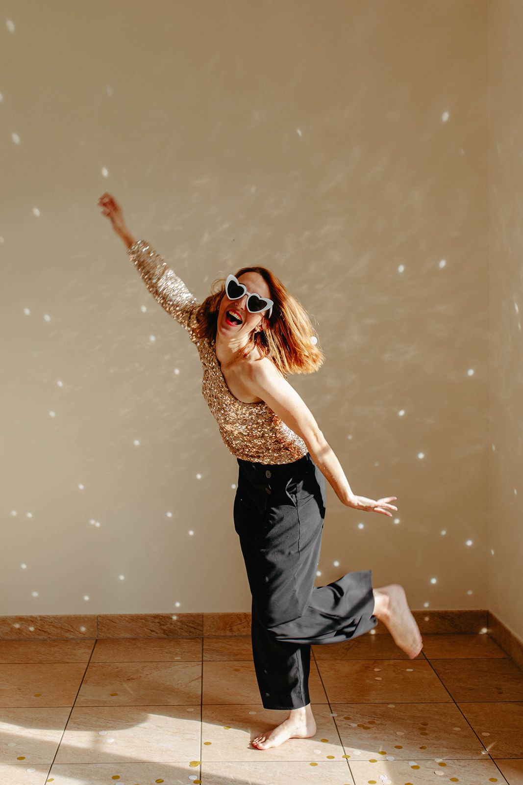 A joyful redhead woman in a sequin top and black pants dancing with confetti falling around her.