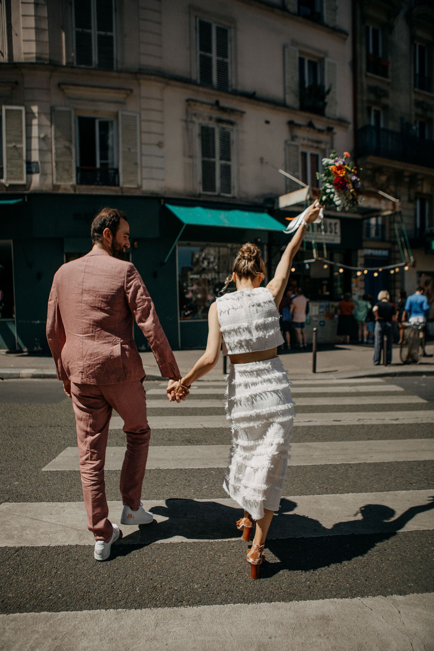 Couple crossing a Parisian street, bride in white fringe dress holding bouquet aloft, groom in pink suit holding hands.