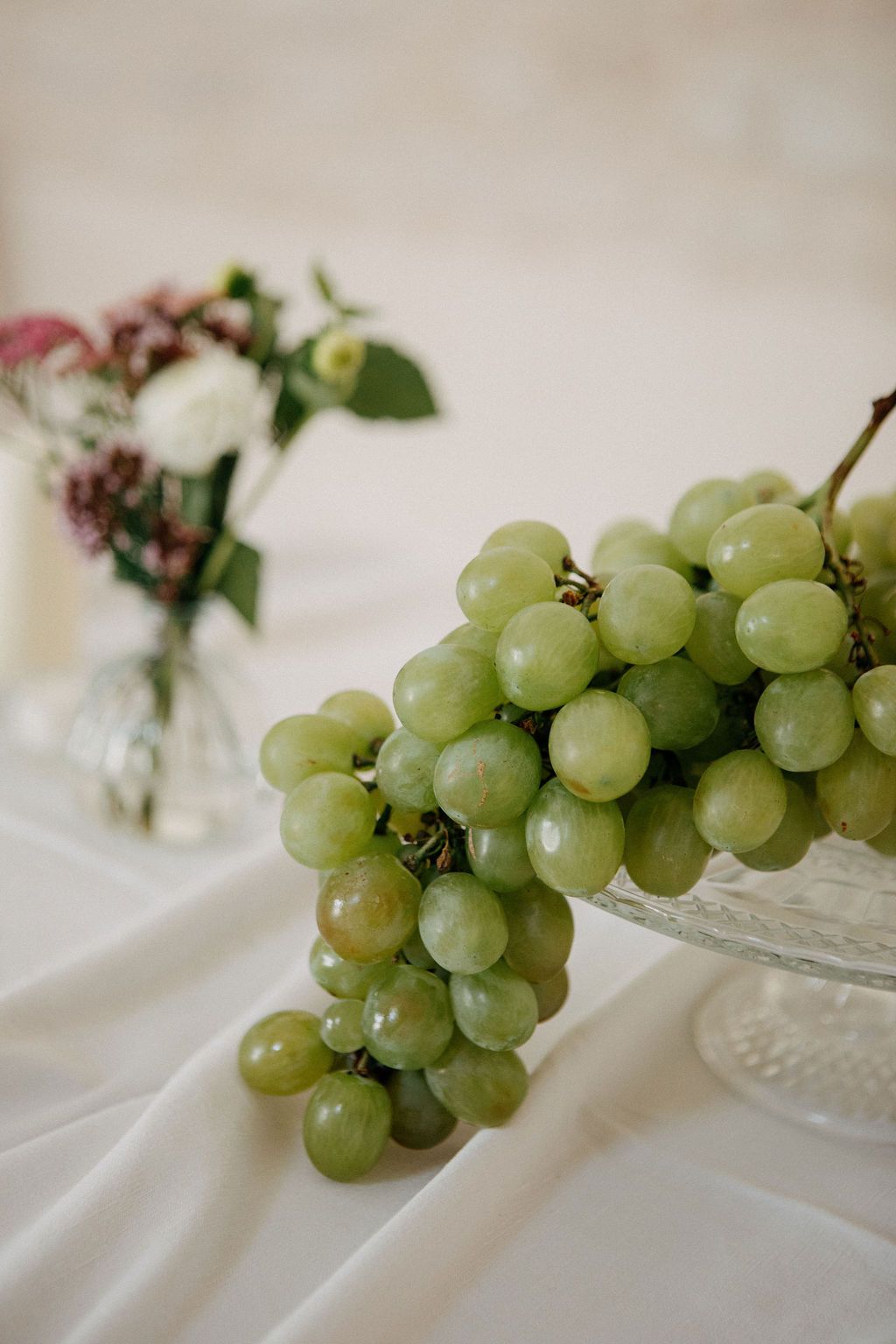 Fresh green grapes on crystal pedestal dish with floral arrangement on white tablecloth at wedding.