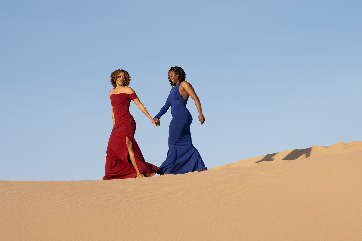 Two figures in contrasting red and blue dresses walking across desert sand dunes.