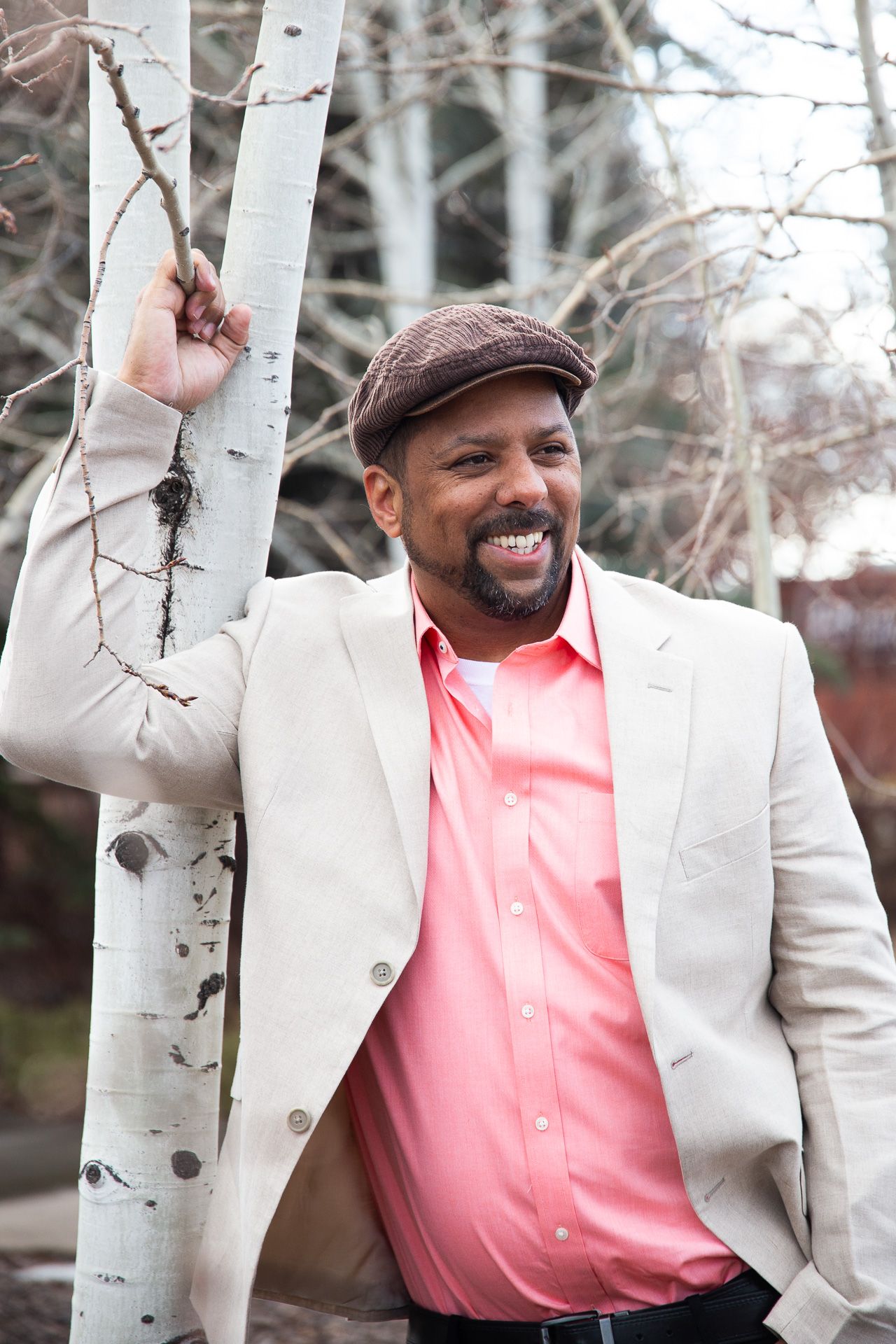 Person in cream suit and coral shirt poses playfully among birch trees in winter setting.
