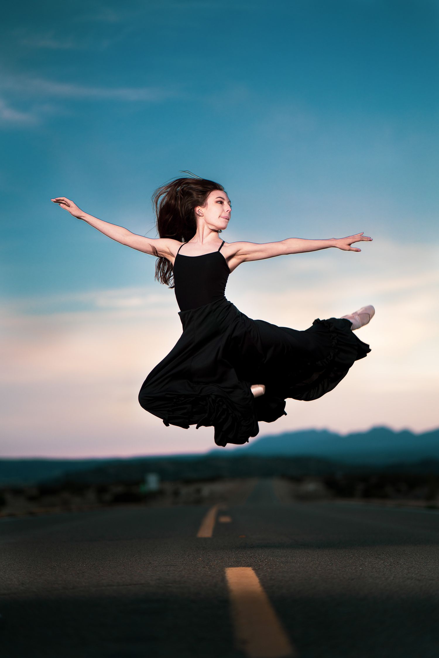 Dancer in black dress leaping gracefully on empty road against blue sky.