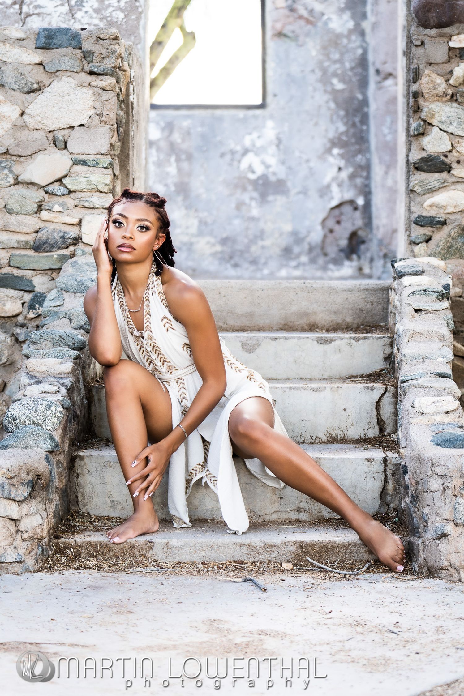 Model in white dress poses elegantly on stone steps in artistic portrait setting.