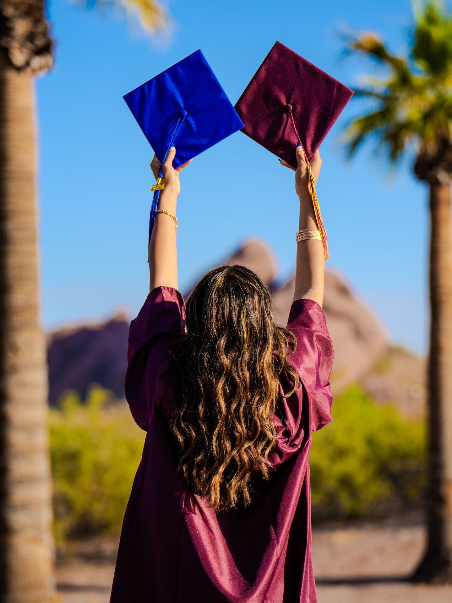 Graduate celebrating outdoors holding up blue diploma cover against palm trees and mountains in background.