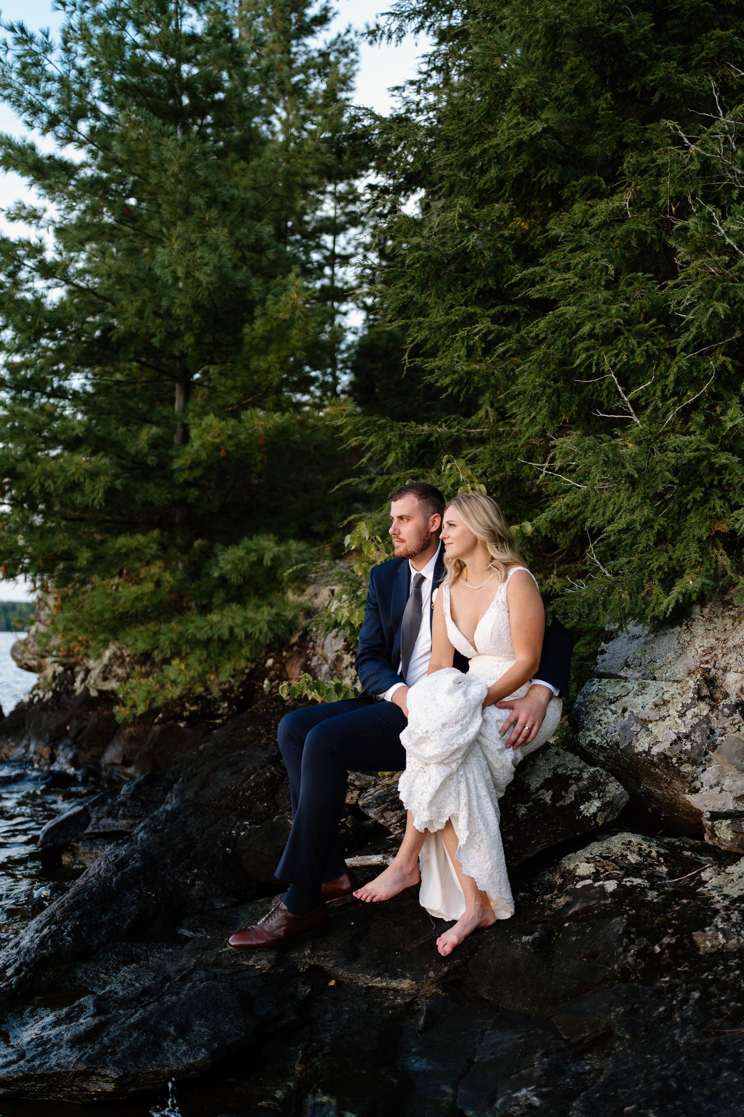 A bride and groom sit together on lakeside surrounded by evergreen trees during a cottage wedding on Lake Joseph- Alexis Anne Photpgraphy