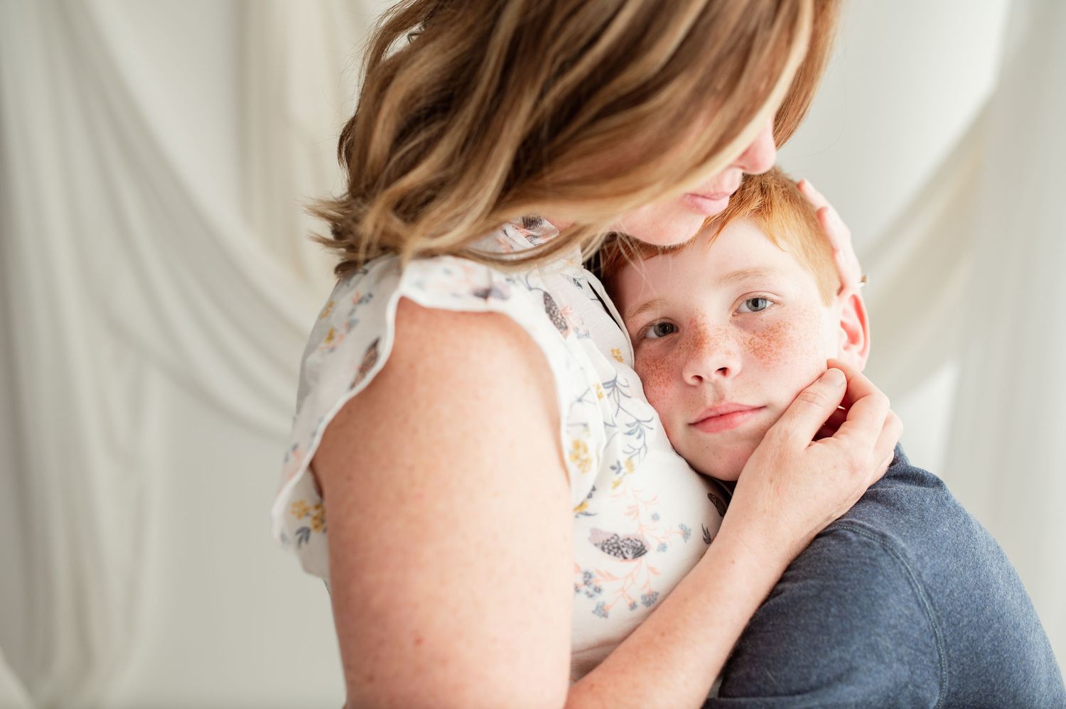 A mother with brown hair tenderly embraces her young red-haired son against a soft white draped background.