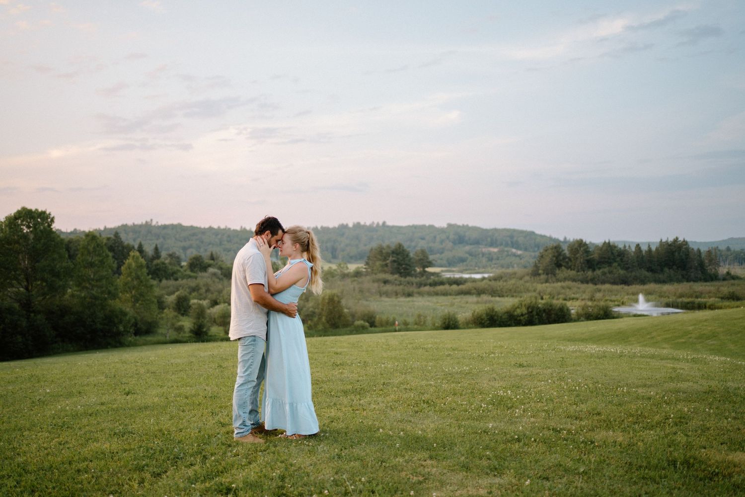 Couple embracing at sunset on a lush green lawn with a scenic countryside backdrop.