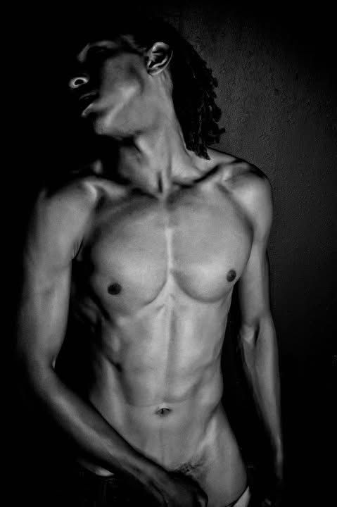 A dramatic black and white artistic nude photograph highlighting muscular physique against a dark background.