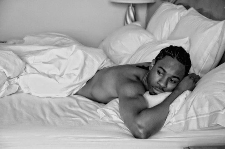 A black and white photograph shows a shirtless figure reclining on white bedding in an intimate pose.