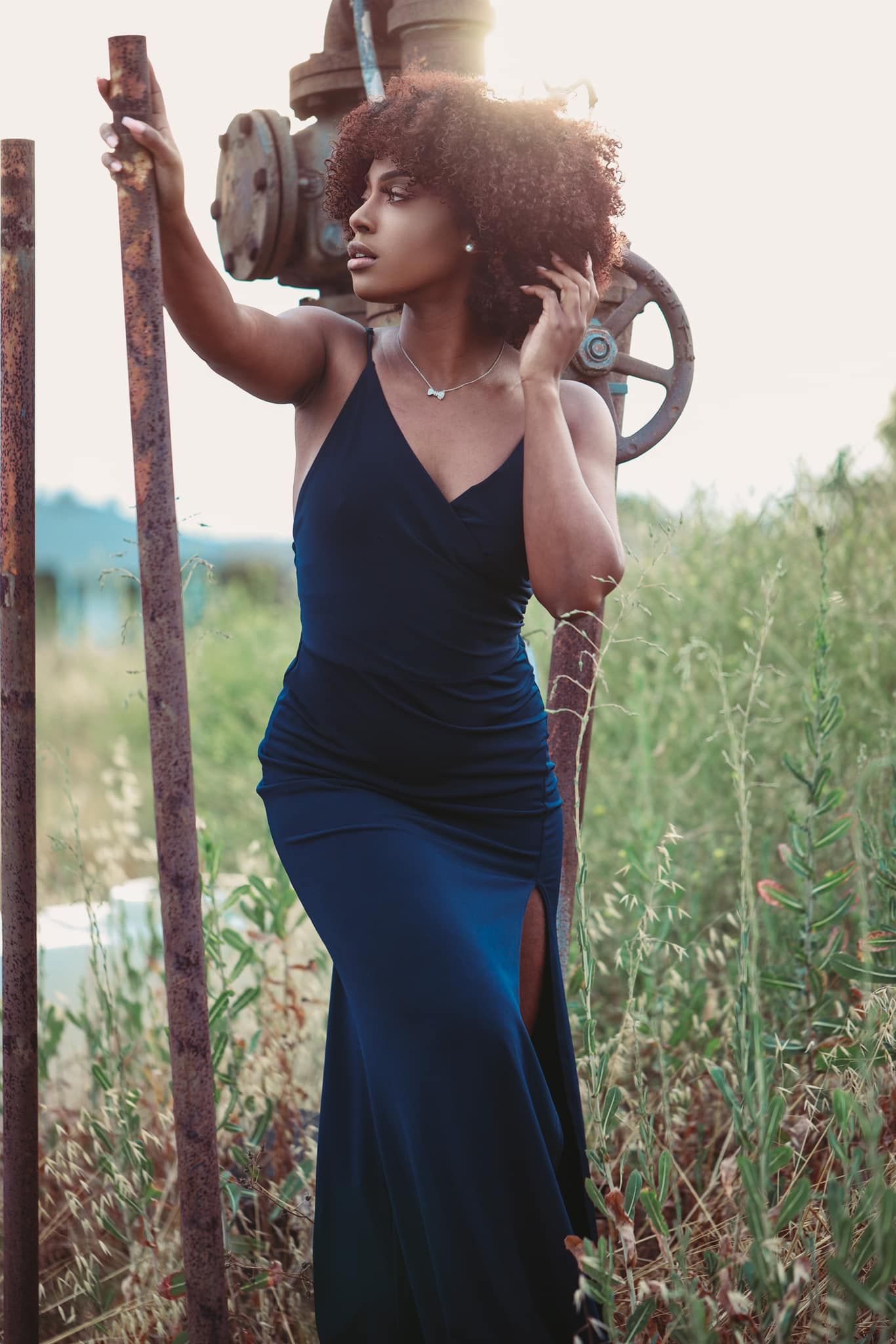 Woman wearing dark elegant dress poses by metal machinery in golden sunlight.