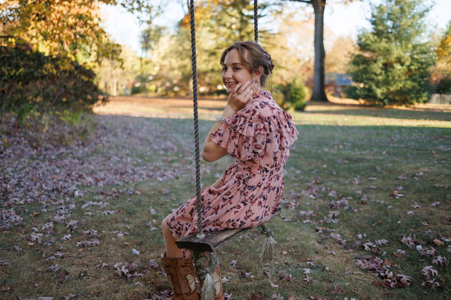 A high school senior girl in a floral dress sits in a grassy outdoor setting during autumn while interacting with a small brown dog.