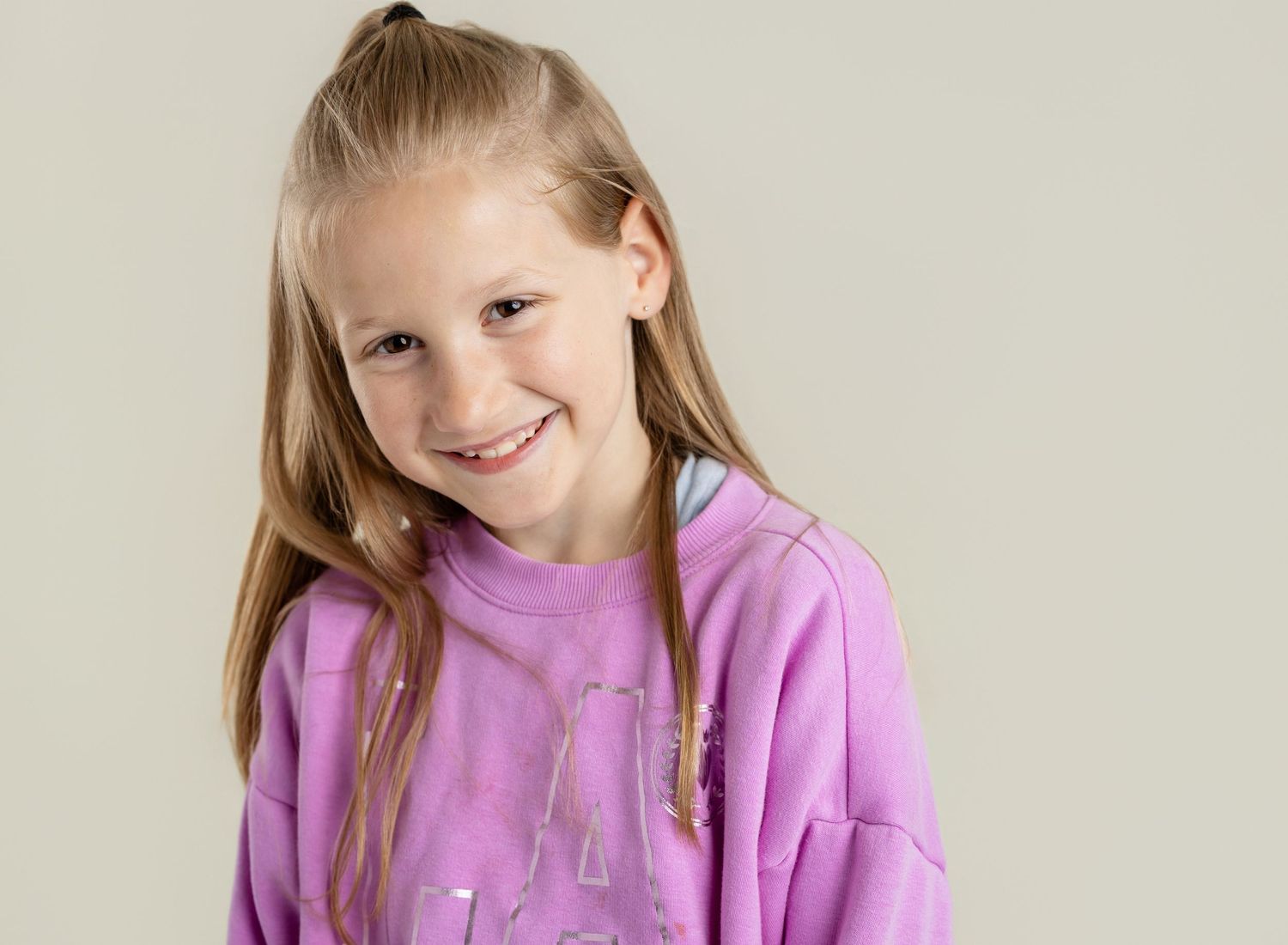 Smiling young girl with blonde hair wearing a pink sweatshirt, posing against a light gray background.