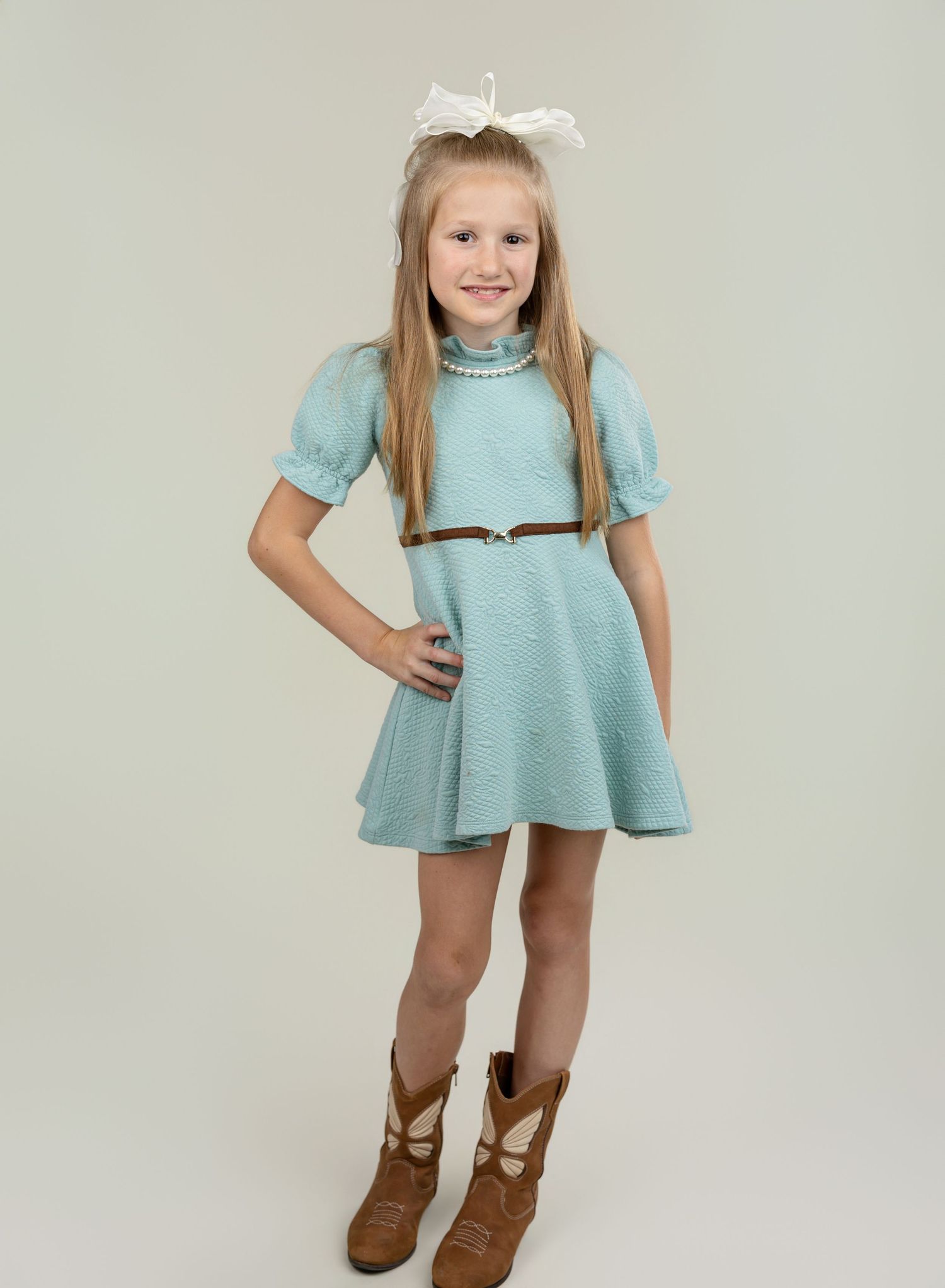 Young girl in mint green belted dress with pearl necklace, brown boots, and white bow hair accessory.