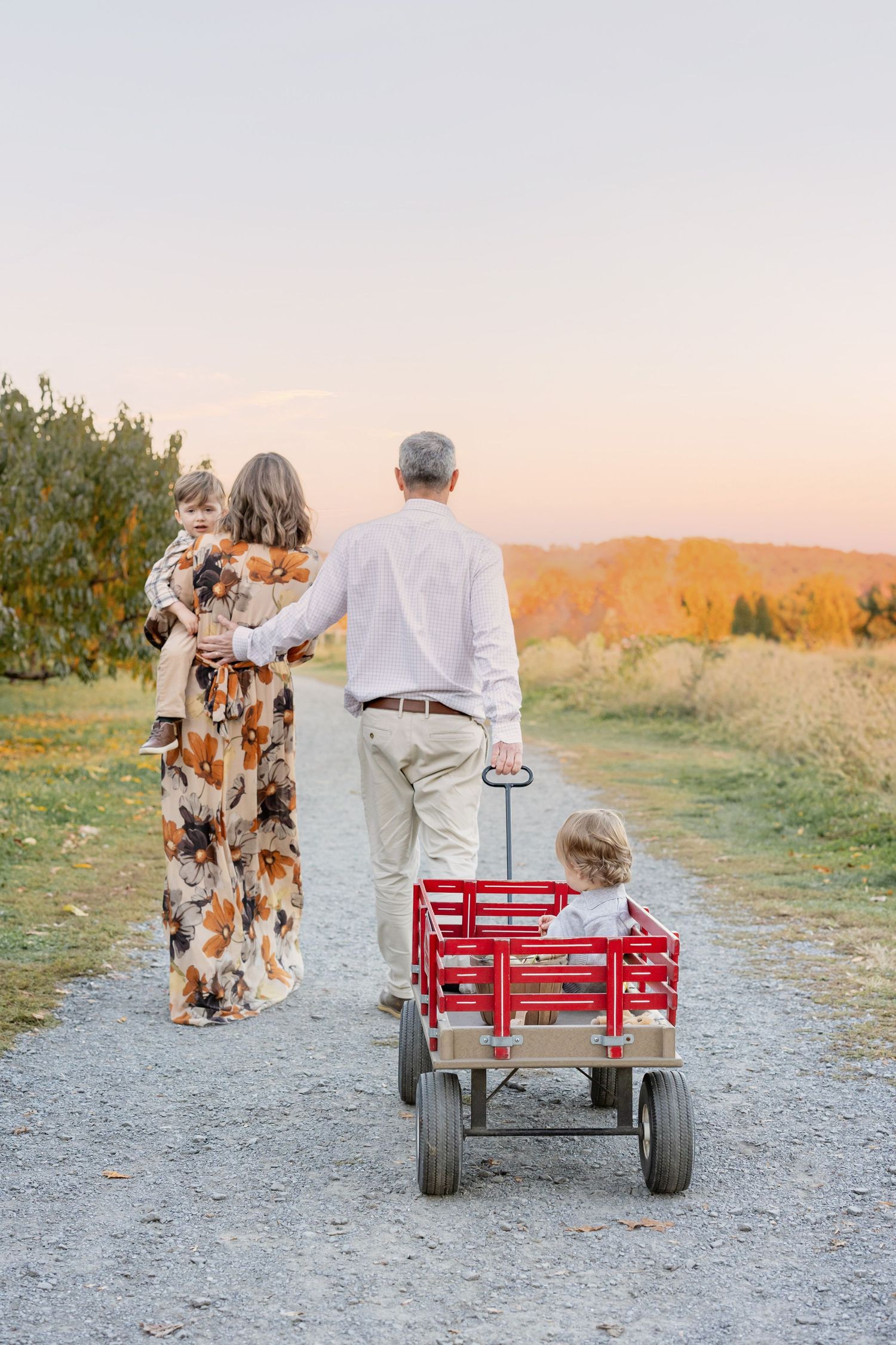 Family walking on gravel path at sunset, parents holding children, pulling red wagon in autumn countryside.