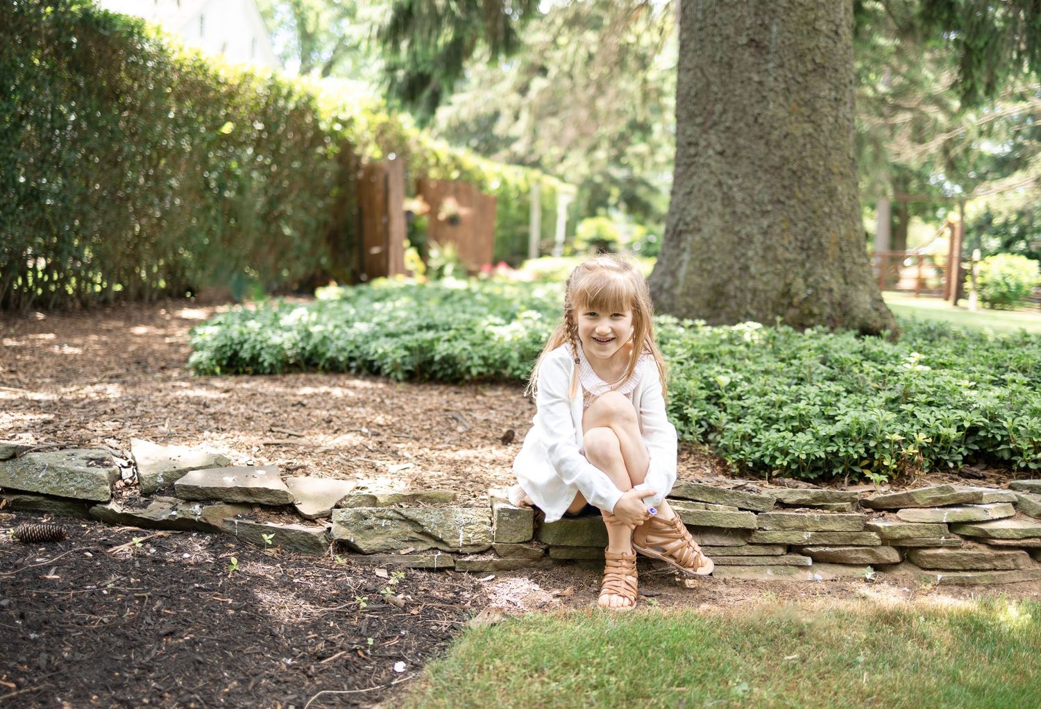A small child in a pink dress sits on a stone retaining wall surrounded by lush garden landscaping at White Chimneys Estate in Gap, PA.