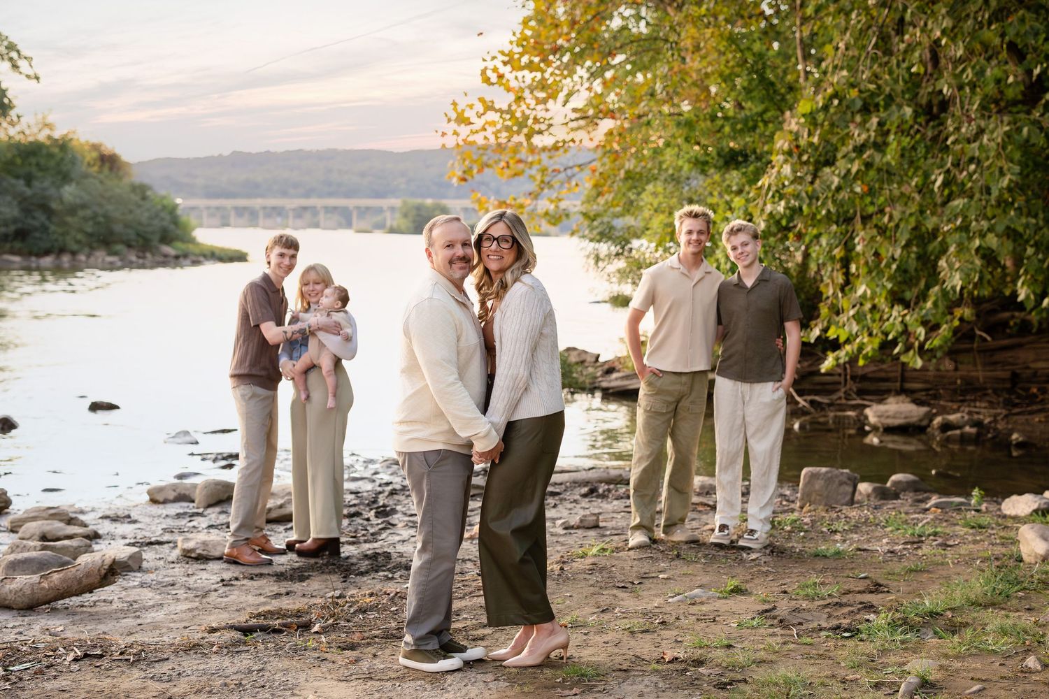 A group stands along a rocky riverbank during golden hour with autumn trees and water in the background.