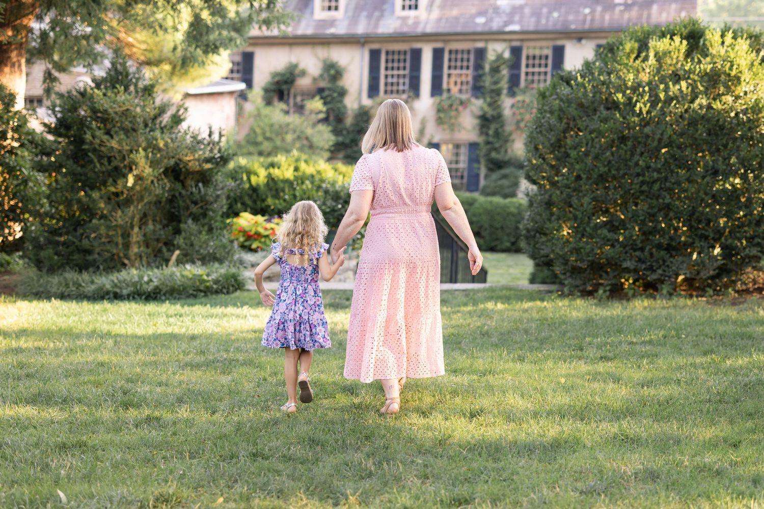 A woman and young girl in dresses walk hand-in-hand across a lush green lawn near a historic stone home.
