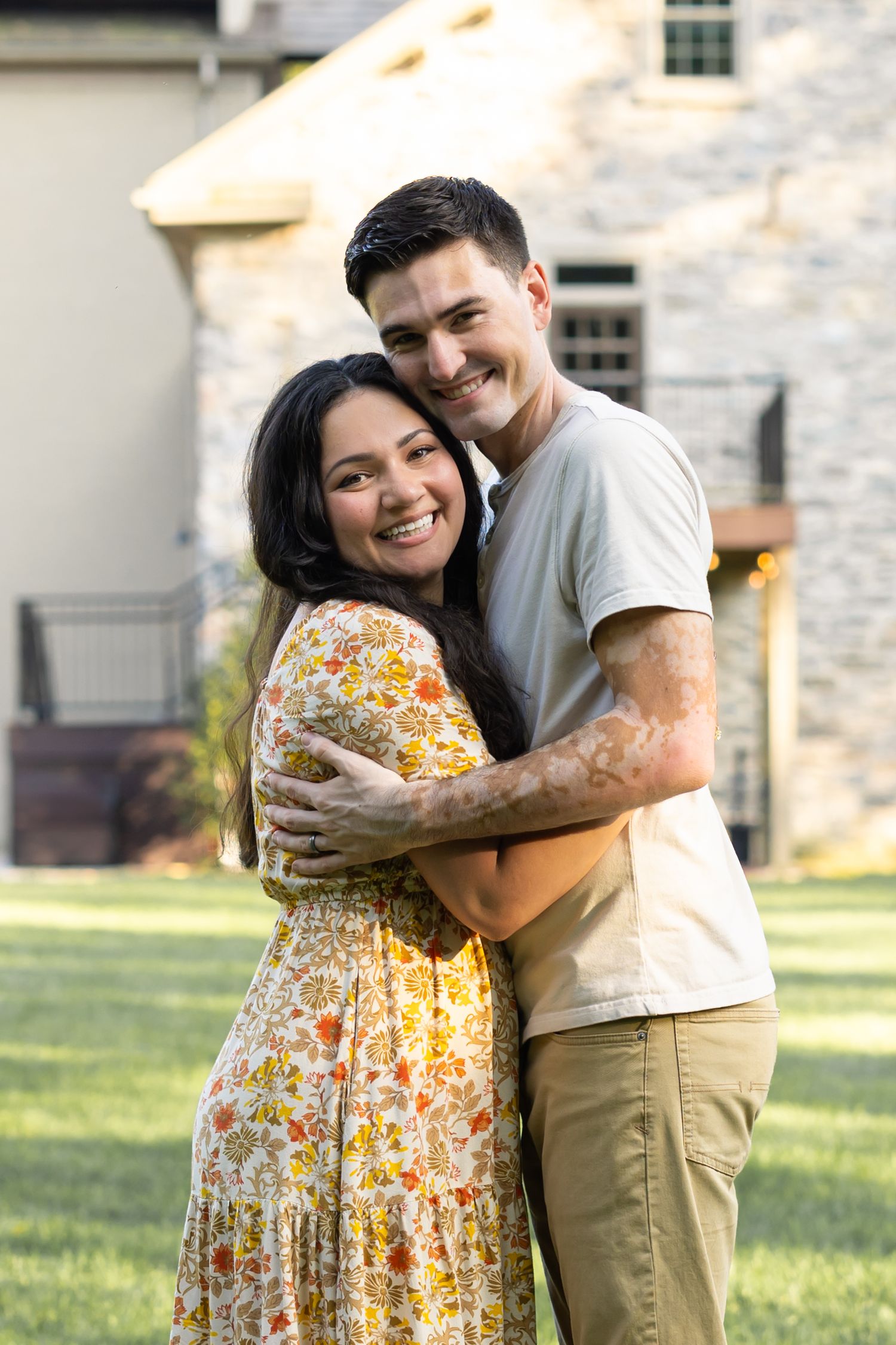 A happy couple embraces for outdoor family photos on a sunny day in Lancaster, PA, with her wearing a floral dress and him in casual attire.