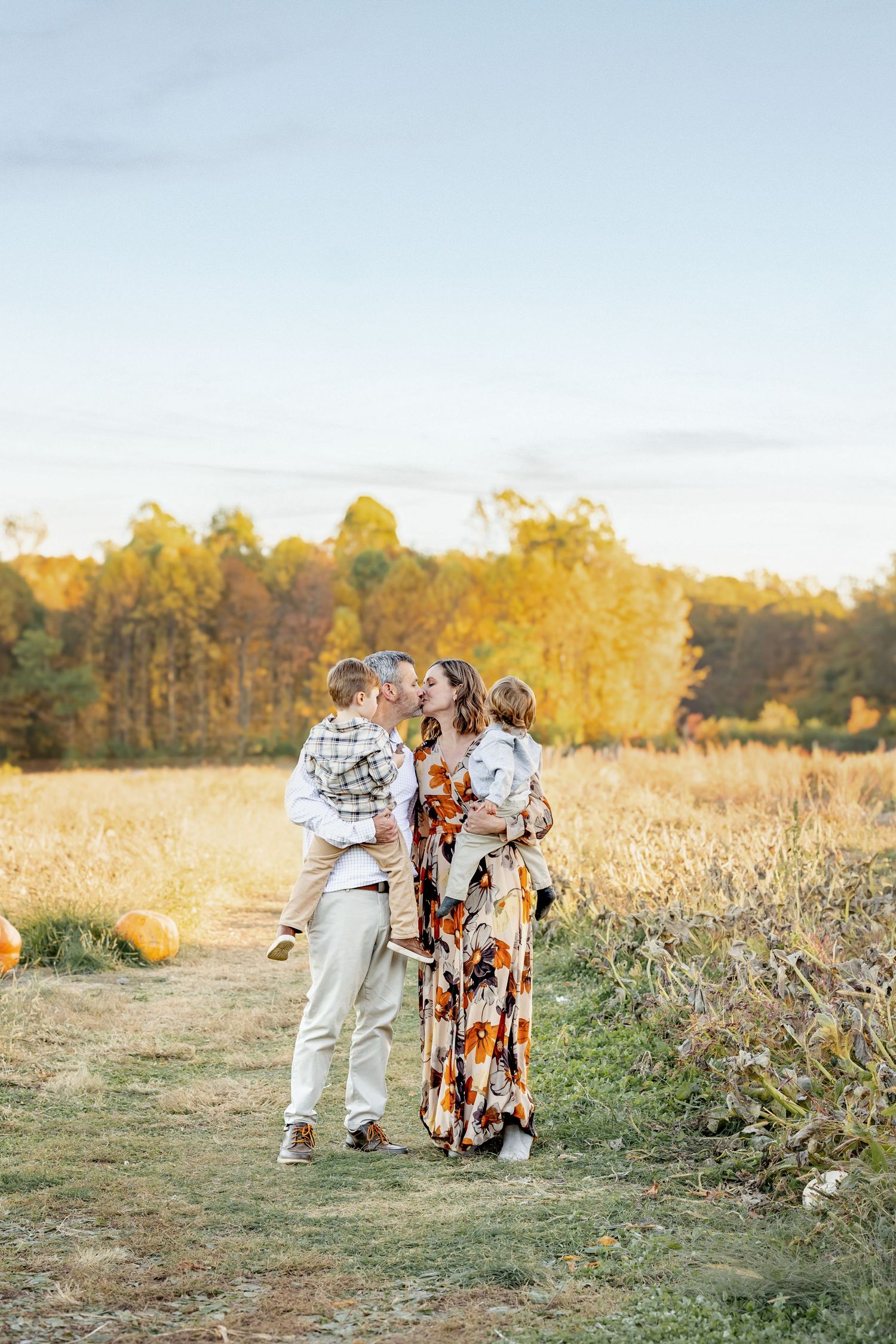 Family sharing kisses in an autumn field, wearing fall colors, with golden trees and pumpkins in the background.