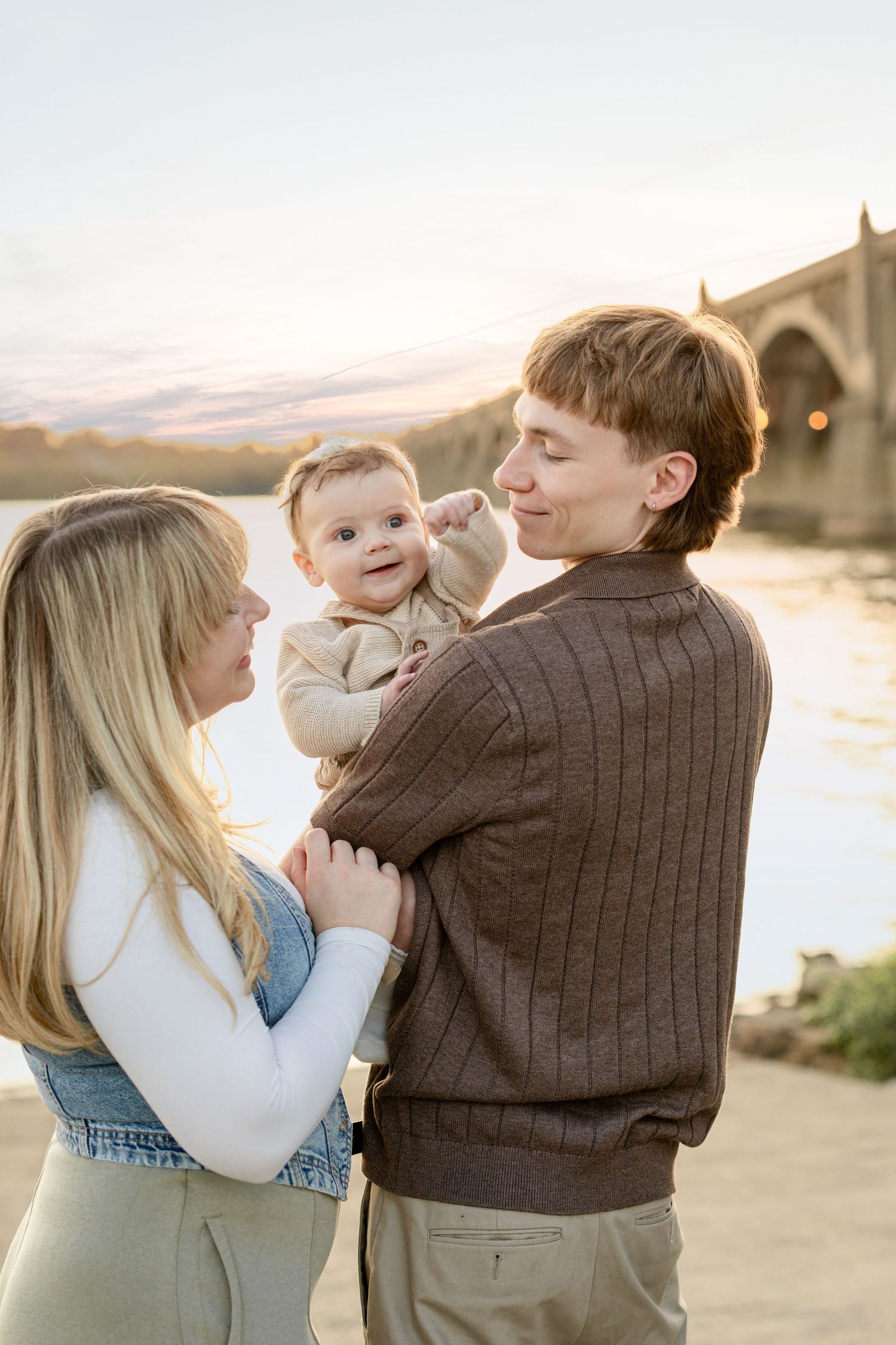 Young family with baby by a river at sunset, parents smiling at infant near a bridge backdrop.