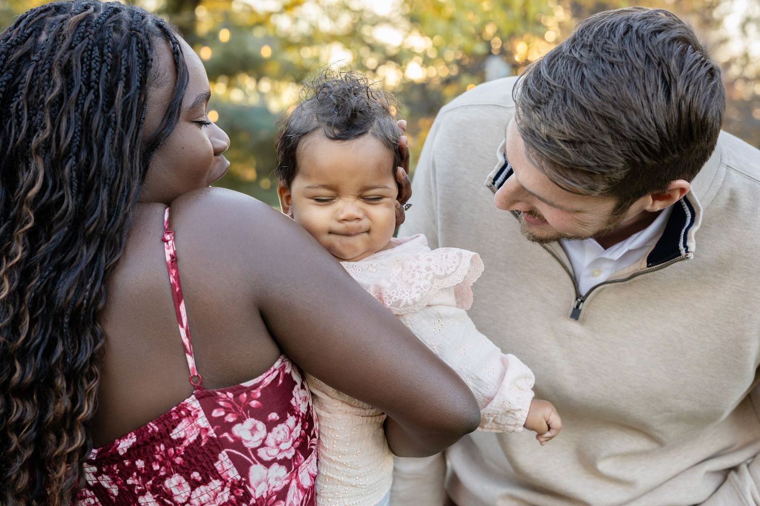 A loving interracial family smiles at their adorable baby girl outdoors in a warm, sunlit park setting.