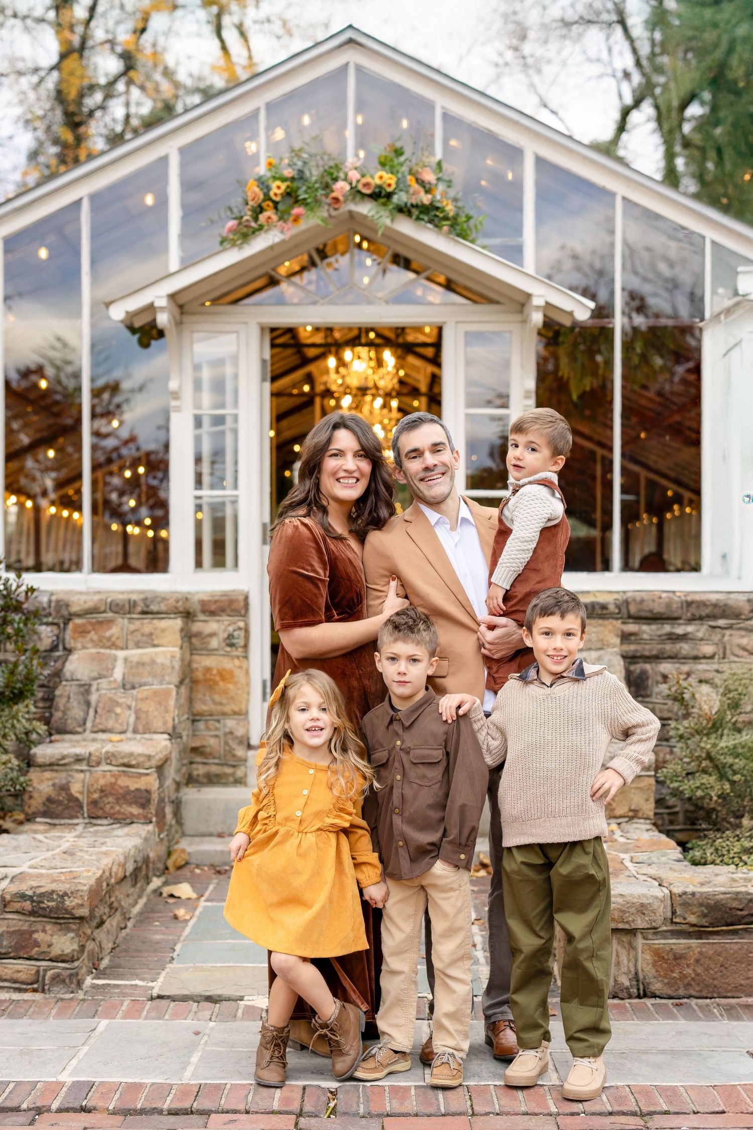 Family of six posing outdoors in front of a decorated greenhouse with string lights and floral accents in autumn.